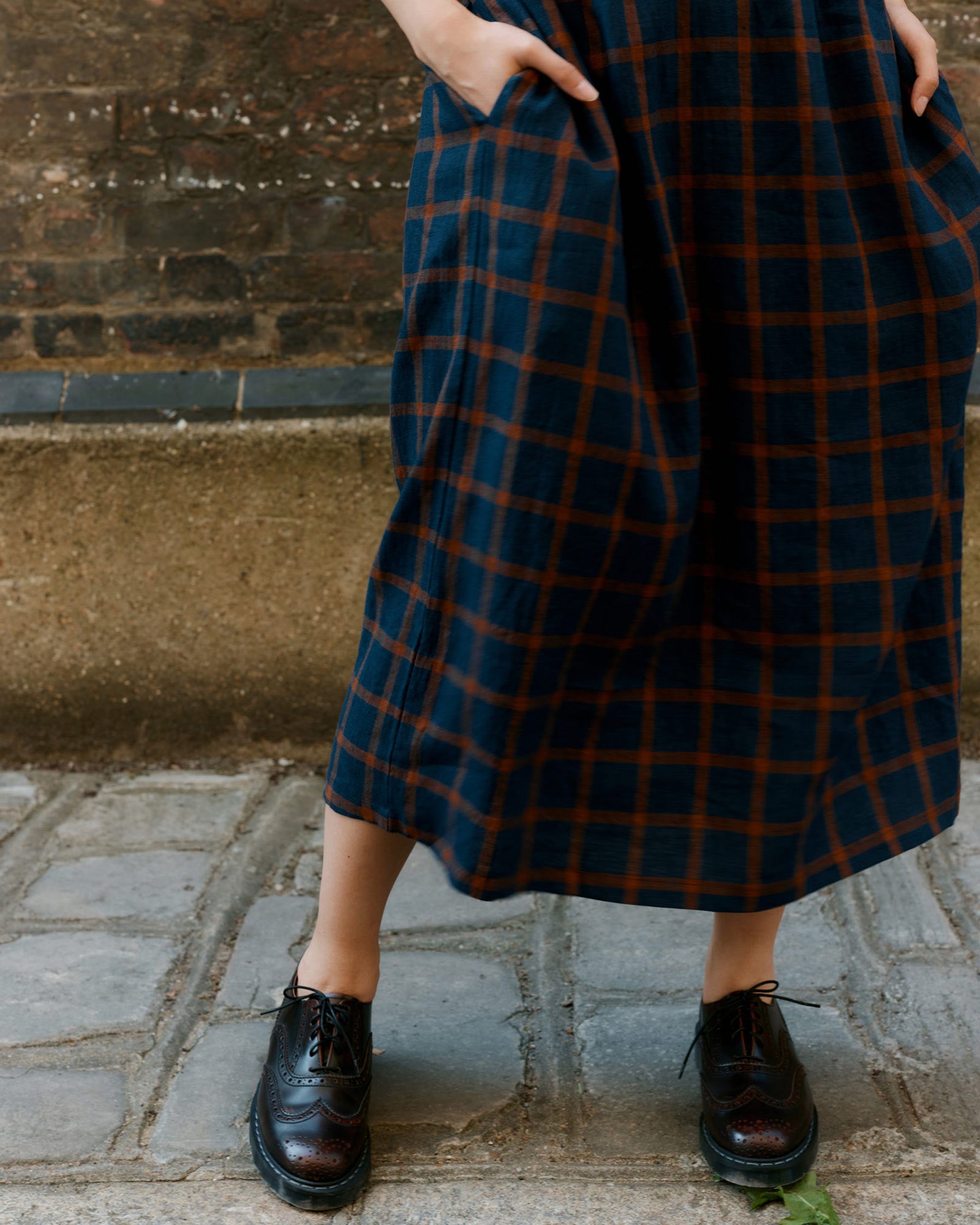 A person stands on a stone path in front of a brick and stone wall, wearing the Pyne and Smith Clothiers No.31 V Neck, Full Length Linen Dress in Montague Windowpane with hands in pockets and black leather lace-up shoes.