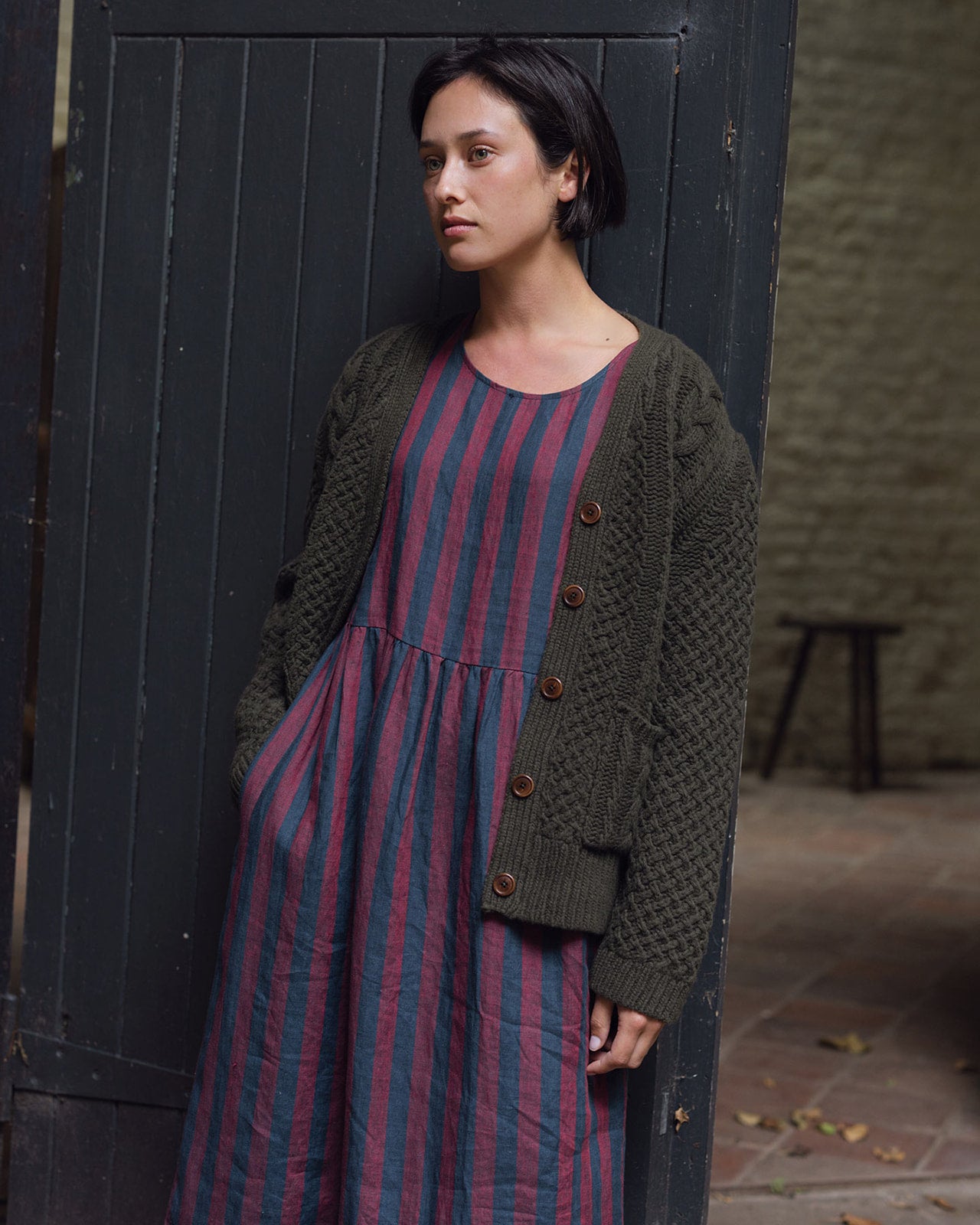 A woman with short dark hair, wearing the Pyne and Smith Clothiers Merino Wool Cable Knit Cardigan with Pockets in Brunswick Green over a loose, striped dress in purple and blue, stands by a dark wooden door looking thoughtful.