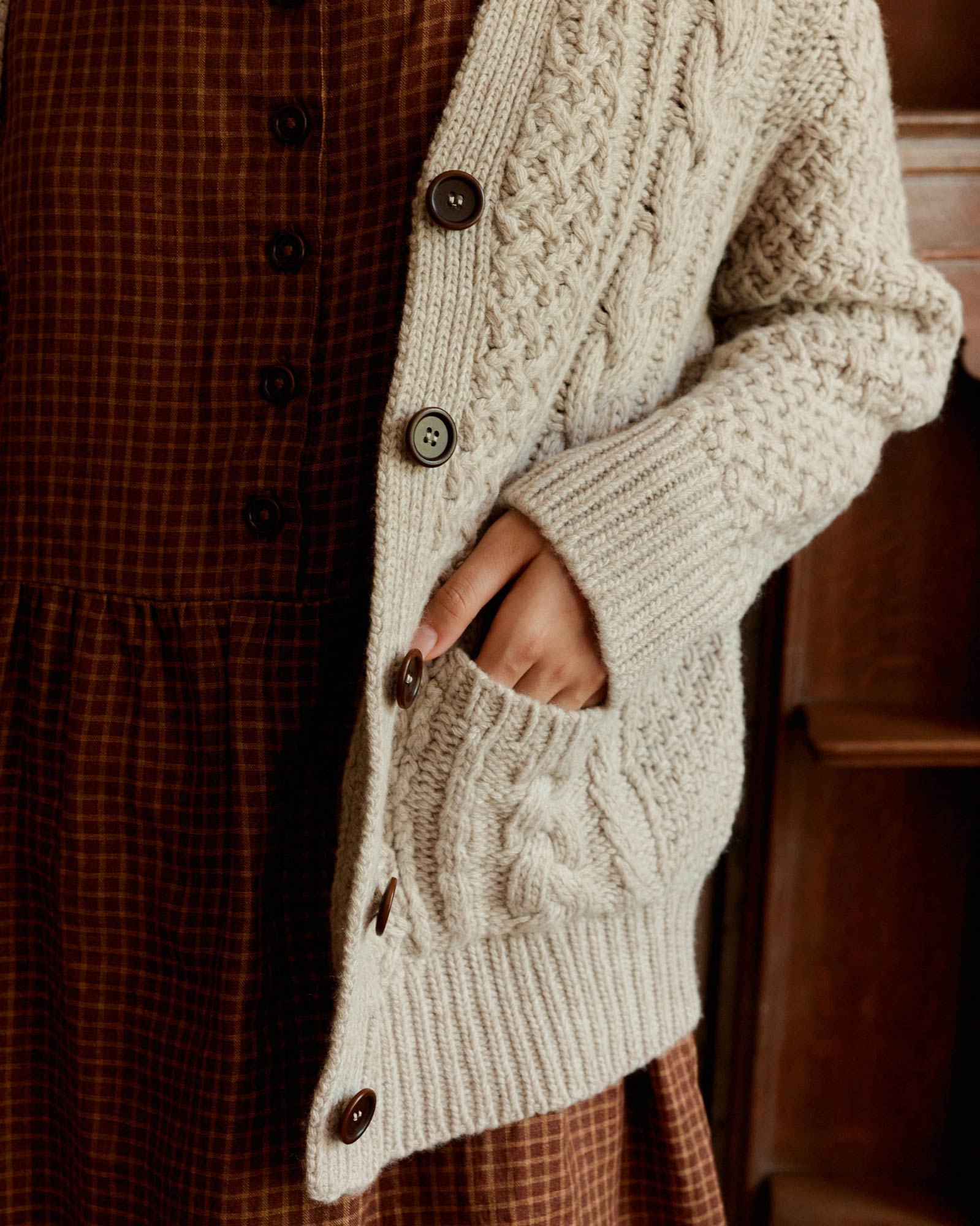 A person wears the Pyne and Smith Clothiers Merino Wool Cable Knit Cardigan with Pockets in Wheaten, partially buttoned, made in Italy, over a brown checkered dress, with one hand tucked into the cardigan pocket.