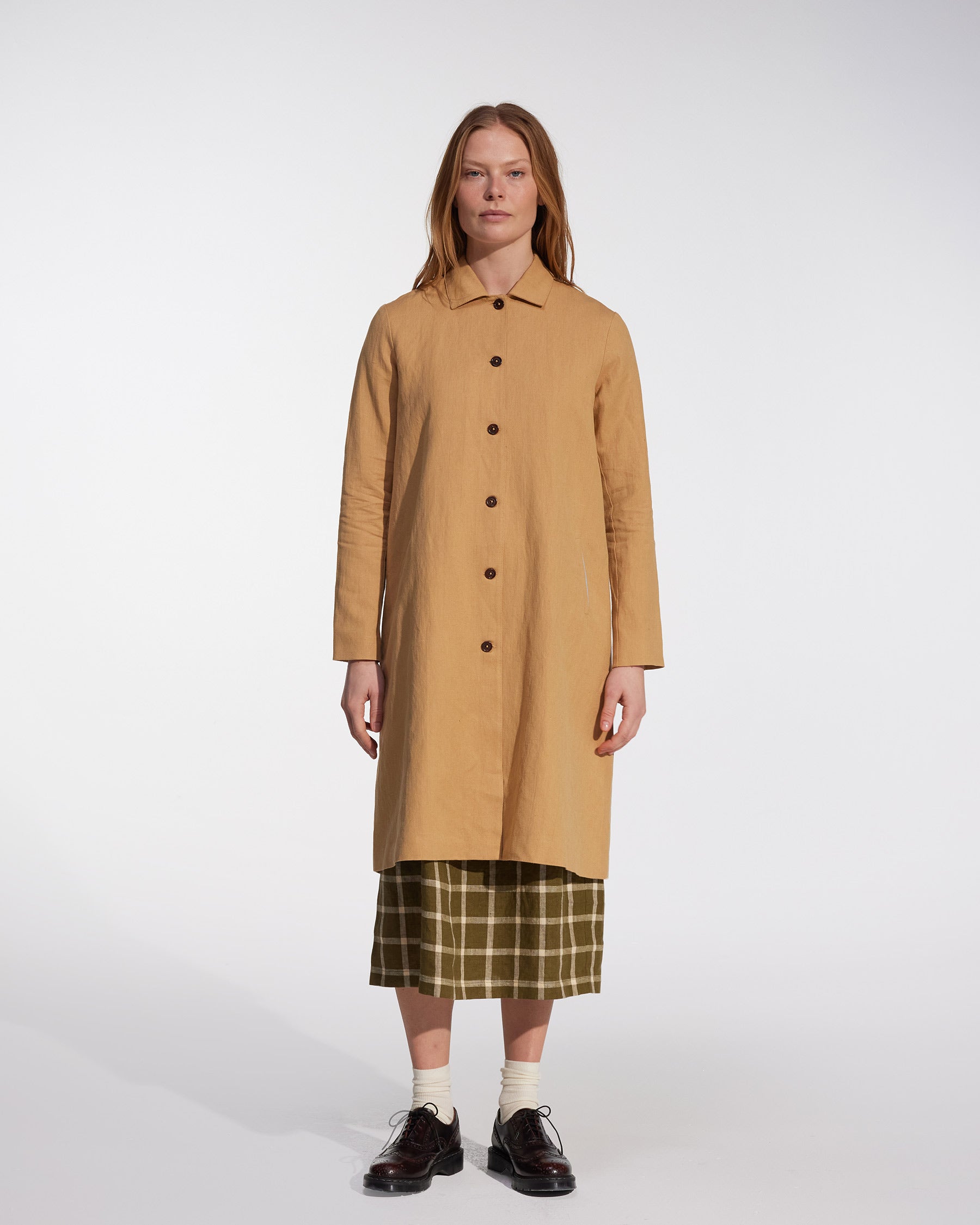 A woman stands in a studio wearing Pyne and Smith Clothiers' Coat No.11 in Lark Tan Cotton Linen Canvas over a green plaid skirt, paired with white socks and dark brown shoes, set against a plain white background.
