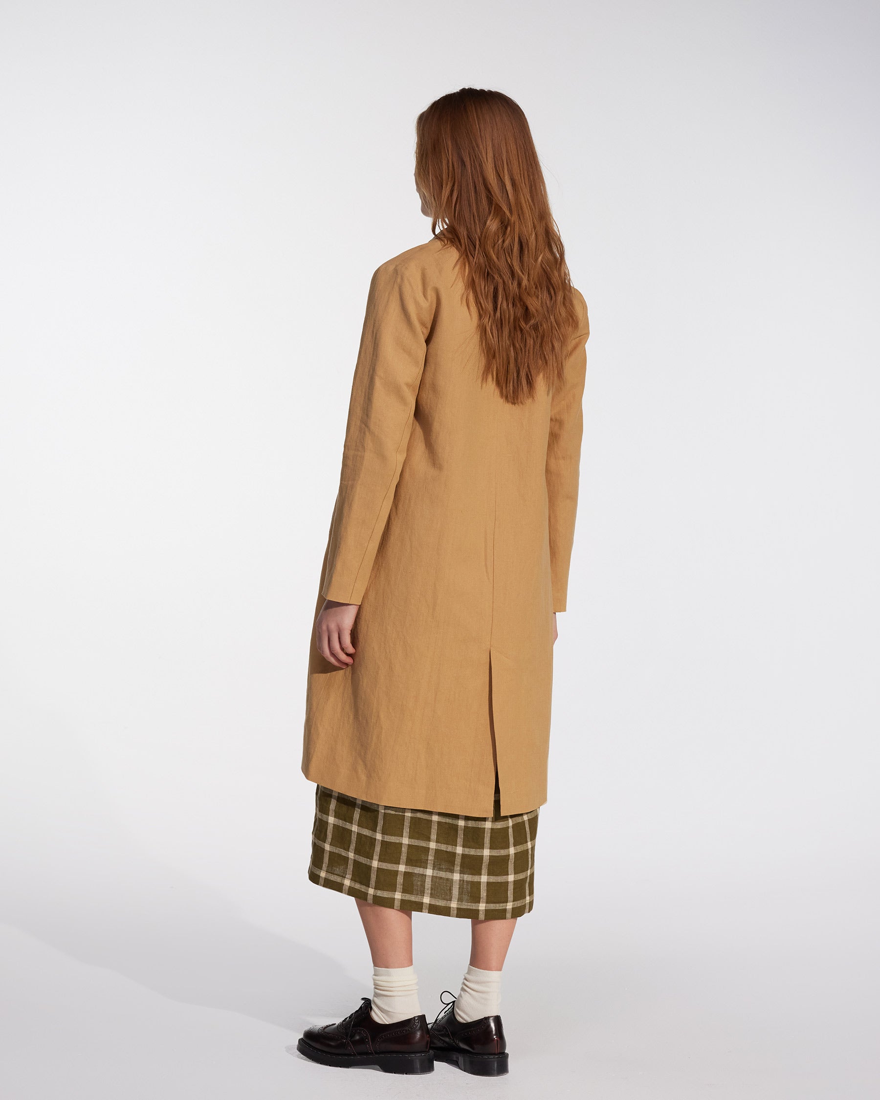 A person with long, wavy hair wears Pyne and Smith Clothiers' Coat No.11 in Lark Tan Cotton Linen Canvas over a green plaid skirt, white socks, and black shoes, standing against a plain white background facing away from the camera.