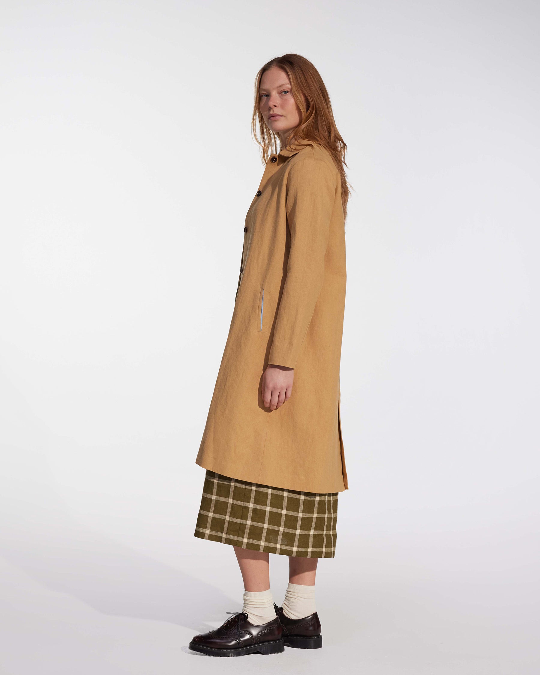 A woman with long reddish-blonde hair stands in profile against a plain white background, wearing the Coat No.11 in Lark Tan Cotton Linen Canvas by Pyne and Smith Clothiers, paired with a plaid skirt, white socks, and dark shoes.