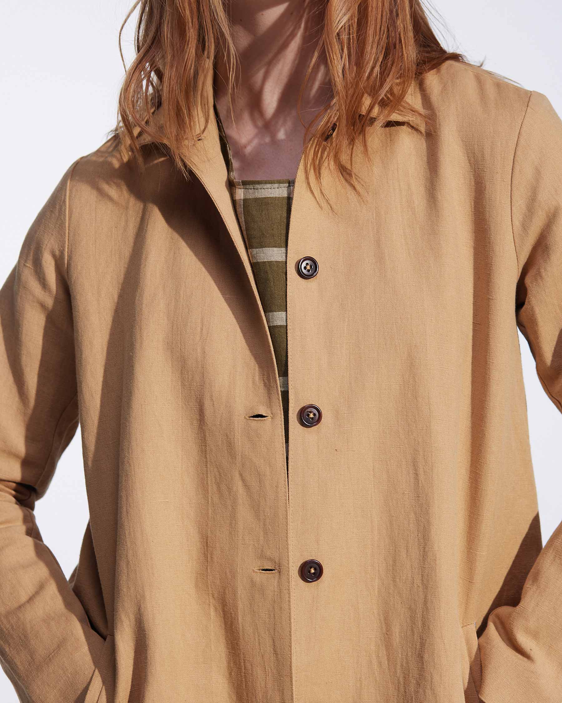 A person with long, light brown hair wears Pyne and Smith Clothiers’ Coat No.11 in Lark Tan Cotton Linen Canvas over a green and beige striped shirt. The image, cropped at the nose and mid-thigh, highlights the chic layered look.