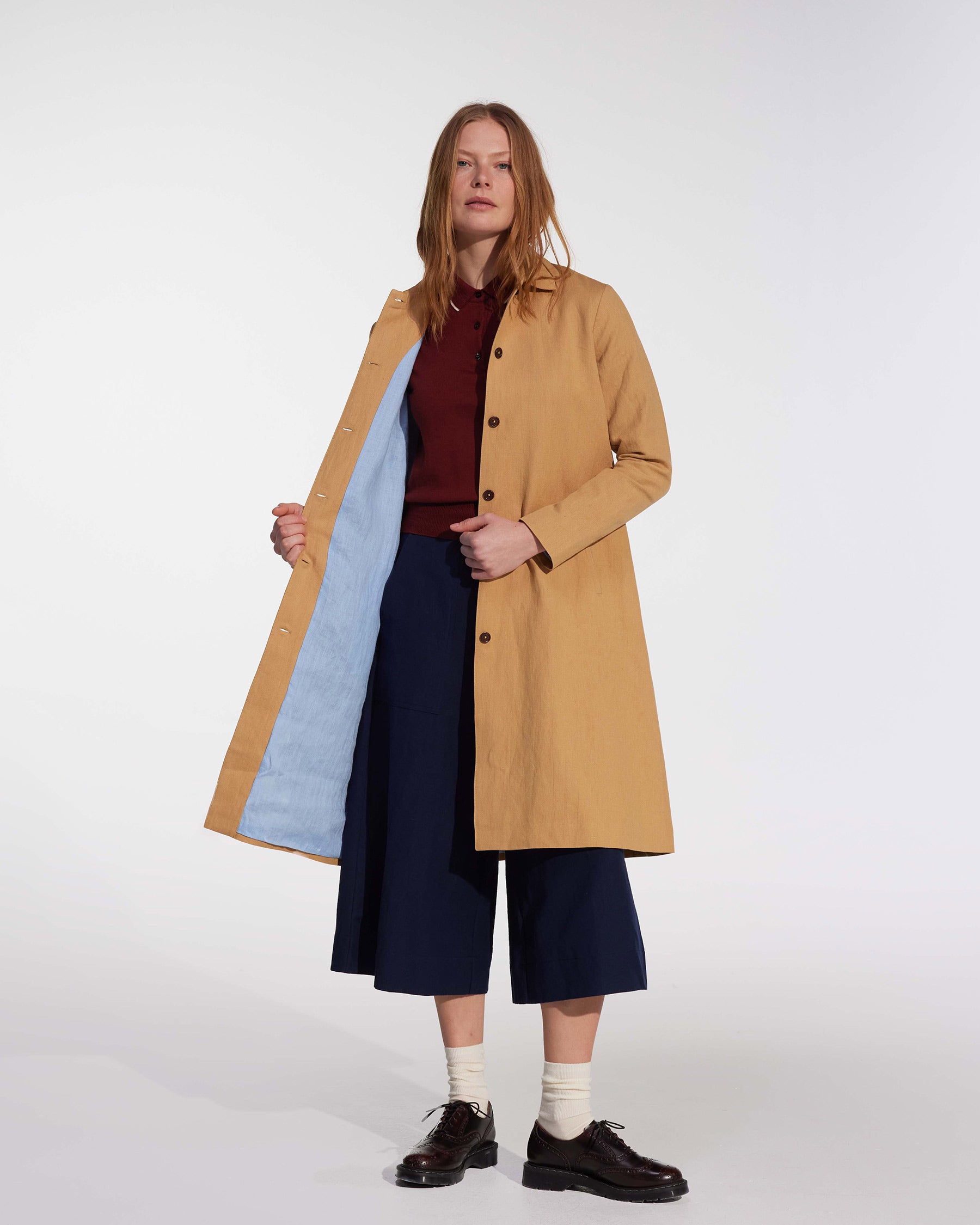 A person stands confidently in the Pyne and Smith Clothiers Coat No.11 in Lark Tan Cotton Linen Canvas, worn over a maroon top, navy wide-leg pants, white socks, and dark shoes against a plain light background.