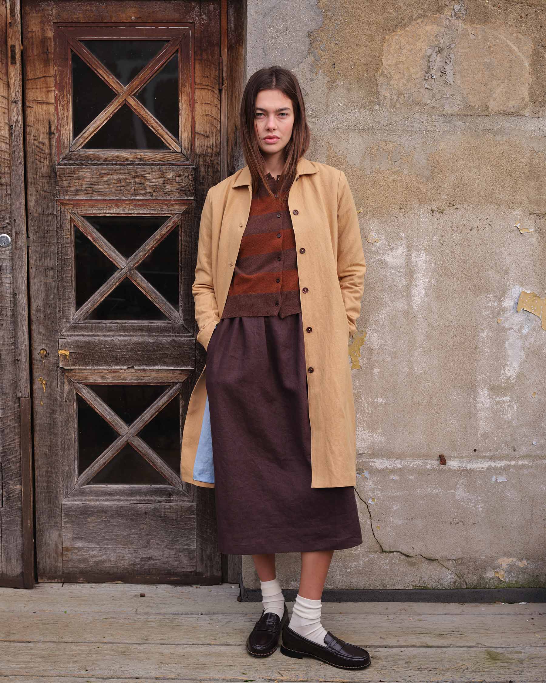 A woman with straight, shoulder-length hair and a neutral expression stands before an old wooden door, wearing the Pyne and Smith Clothiers Coat No.11 in Lark Tan Cotton Linen Canvas over a brown sweater and skirt, with white socks and black loafers.