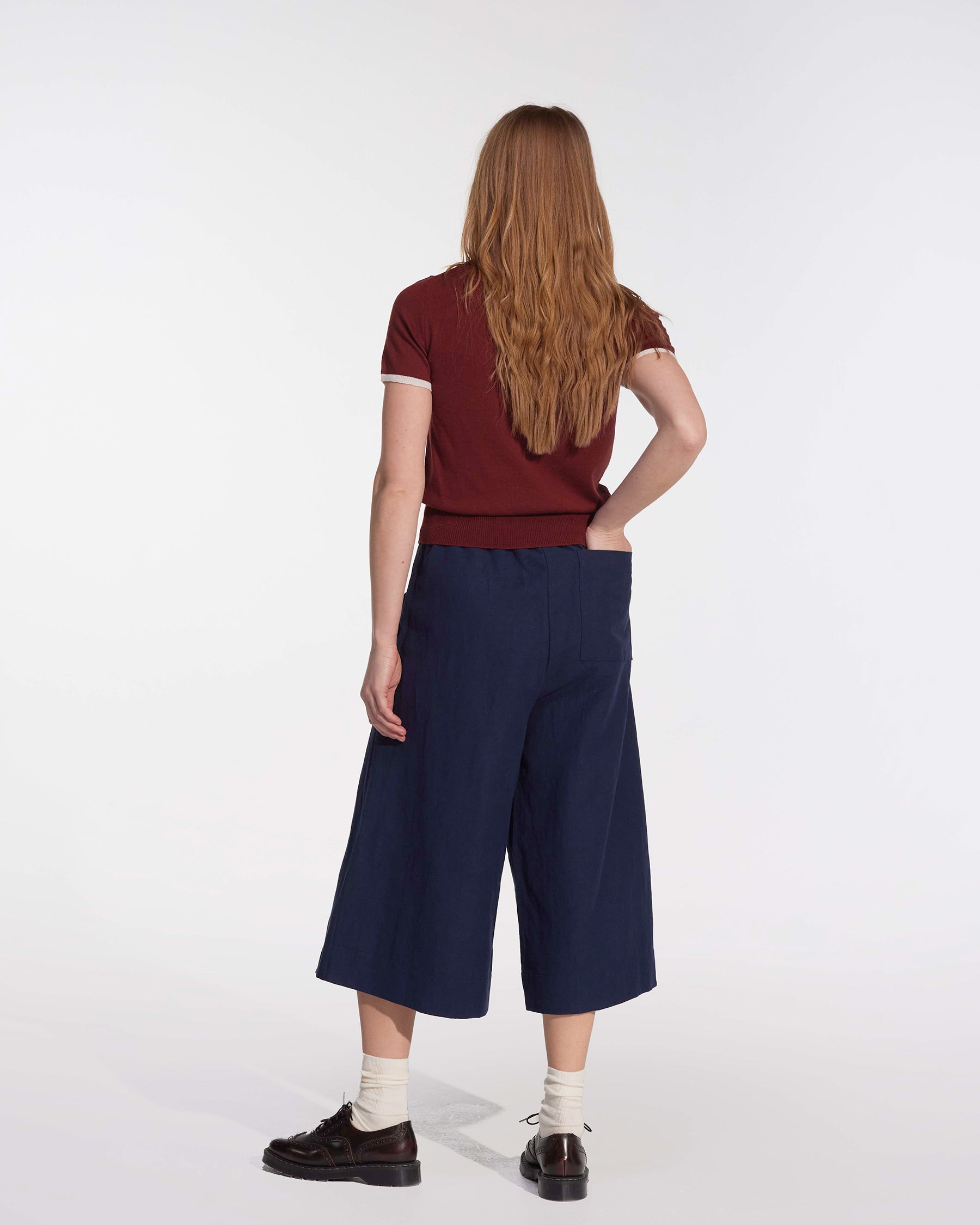 A person with long, straight, reddish-brown hair stands facing away in a maroon shirt and Pyne and Smith Clothiers' Canvas Cropped Trousers No.17 in Ink Blue, with white socks and black shoes against a plain white background.