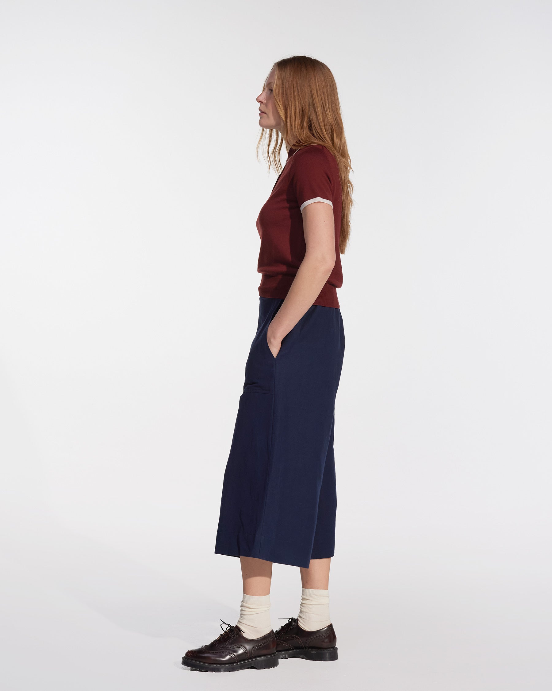 A woman with long red hair stands in profile against a white background, hands in pockets, wearing Pyne and Smith Clothiers' Canvas Cropped Trousers No.17 in Ink Blue with a maroon top, white socks, and dark brown shoes.