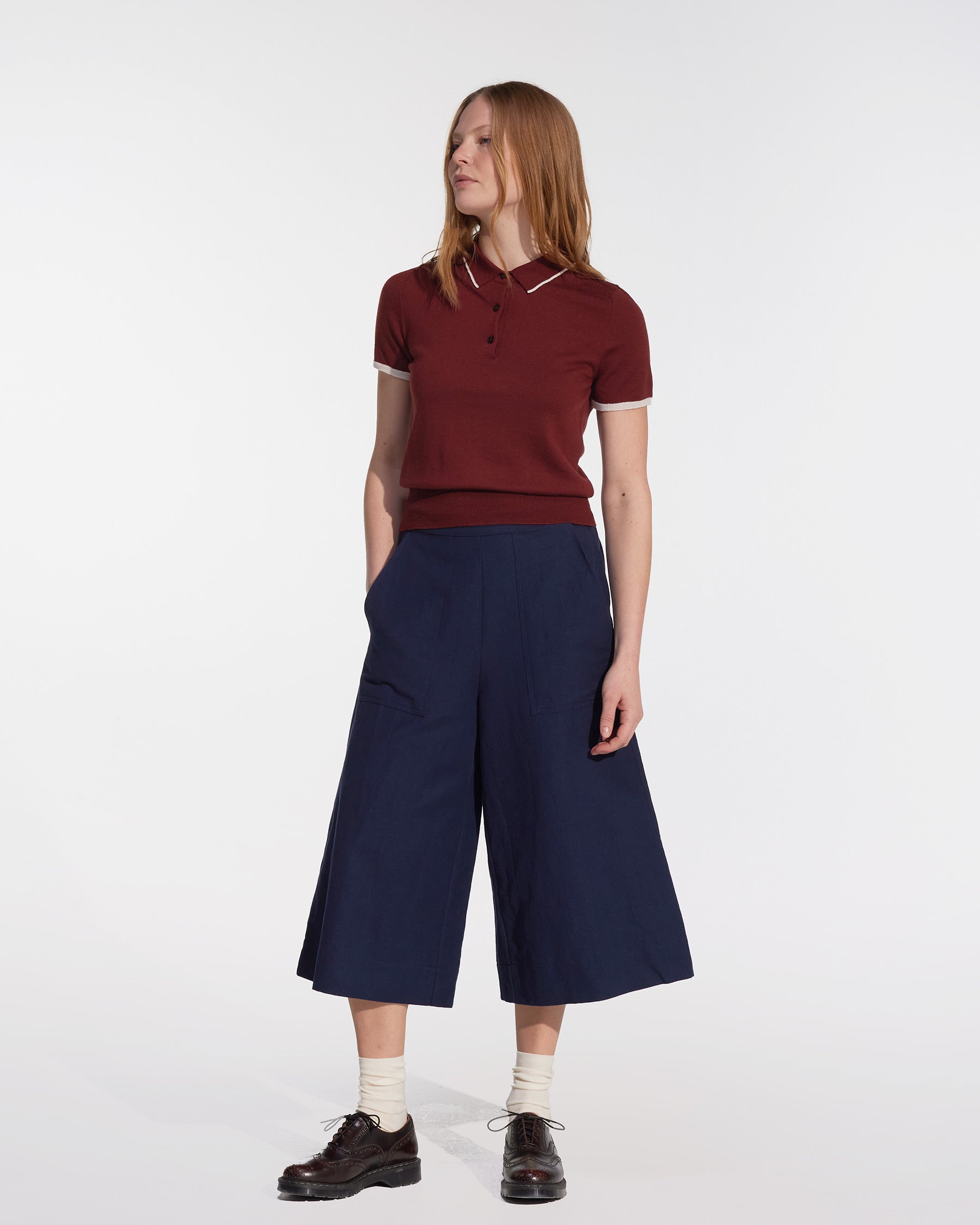 A woman with straight, shoulder-length hair stands against a plain background, wearing a burgundy short-sleeve polo shirt, Pyne and Smith Clothiers Canvas Cropped Trousers No.17 in Ink Blue, white socks, and dark brown shoes. She looks slightly to the side.