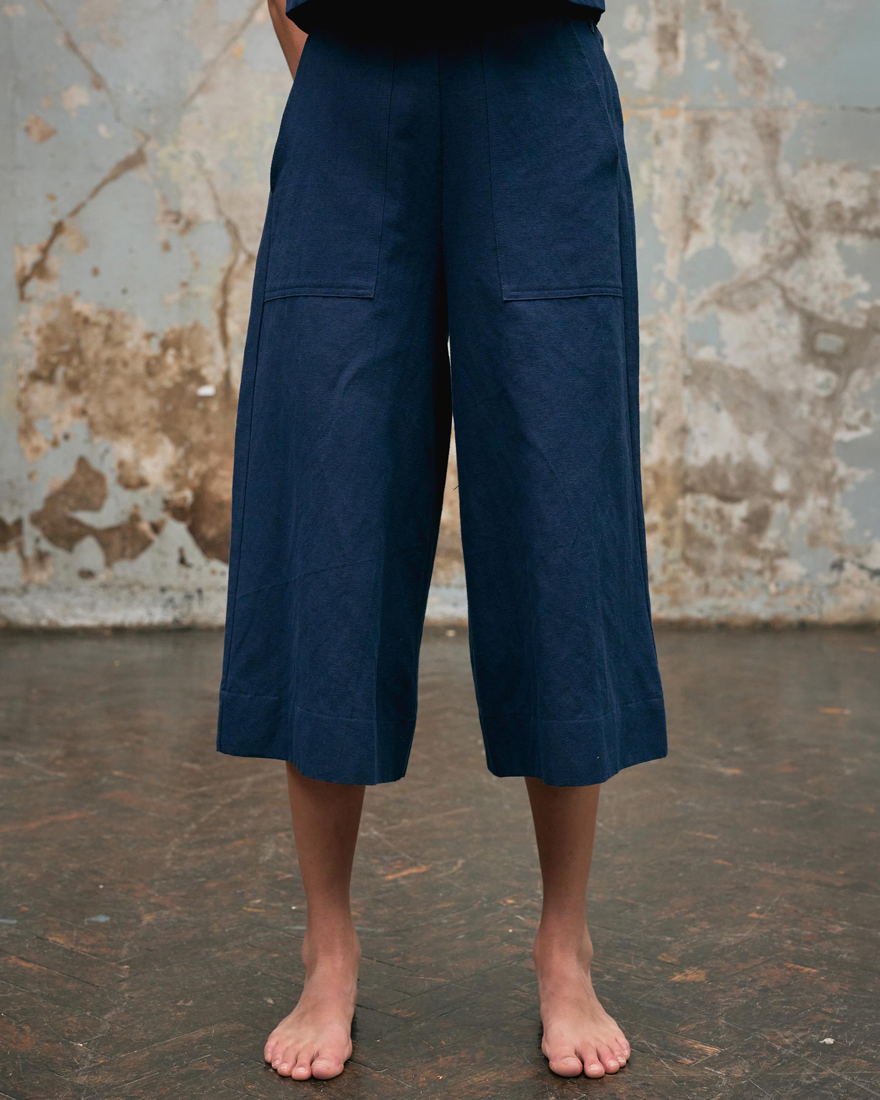 A person stands barefoot on a worn brown floor, wearing Pyne and Smith Clothiers' Canvas Cropped Trousers No.17 in Ink Blue, with a distressed wall and peeling paint in the background. Their upper body is mostly out of frame.
