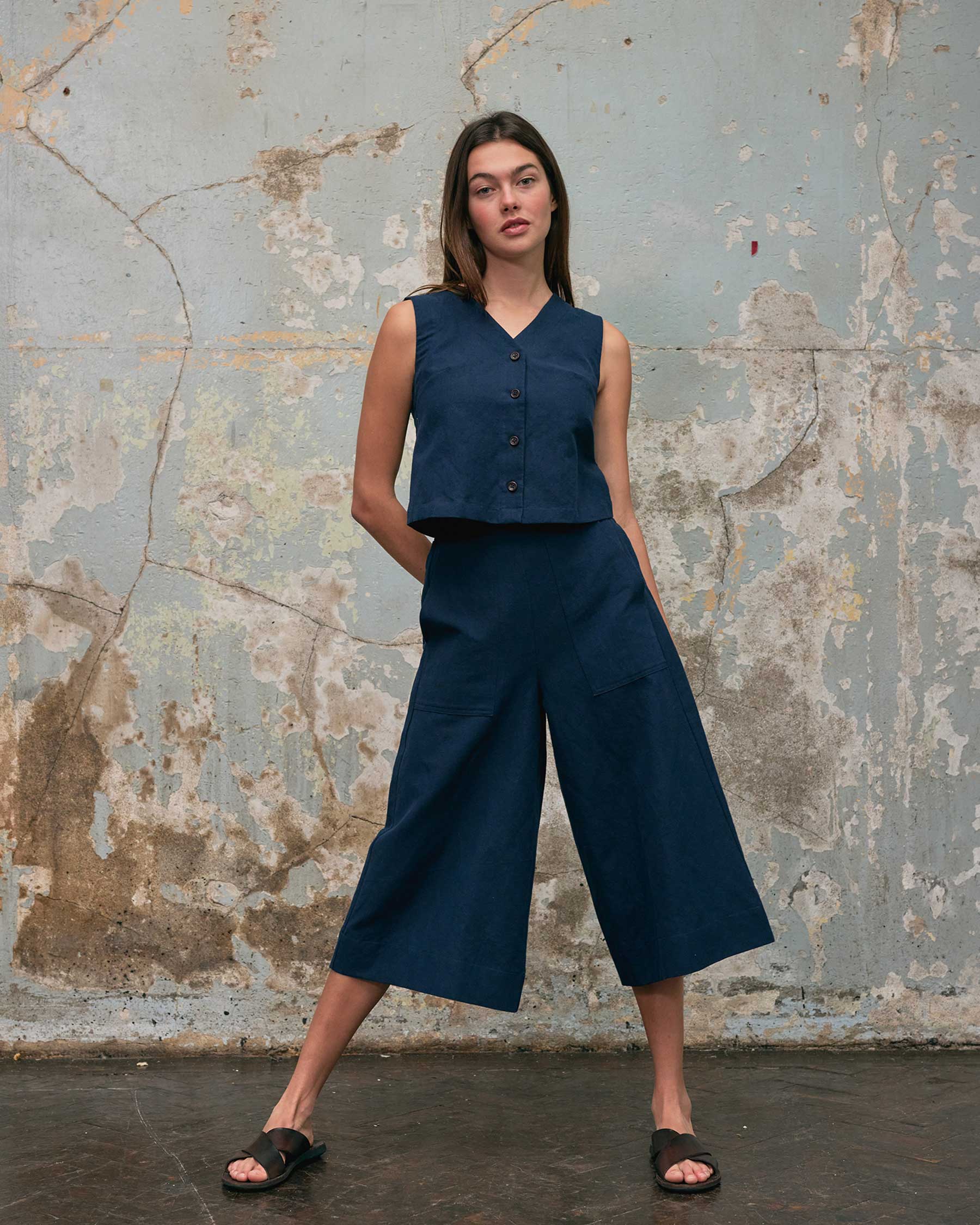 A woman with straight brown hair stands confidently before a textured wall, wearing Pyne and Smith Clothiers' Button up Vest in Ink Blue Canvas with matching wide-leg culottes and black sandals.
