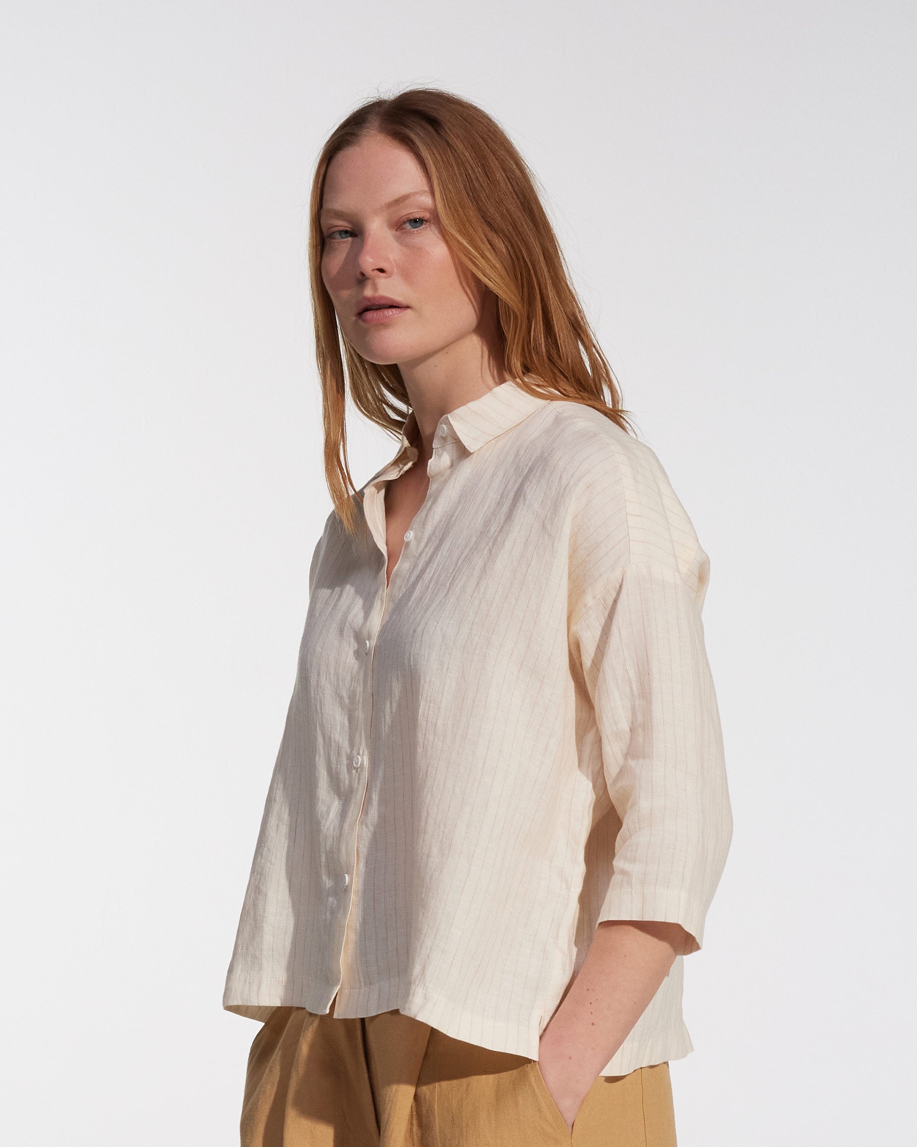 A woman with long reddish-blonde hair wears the Pyne and Smith Clothiers Boxy Cropped Linen Blouse No.22 in Cream Pinstripe and tan pants, standing against a plain white background.