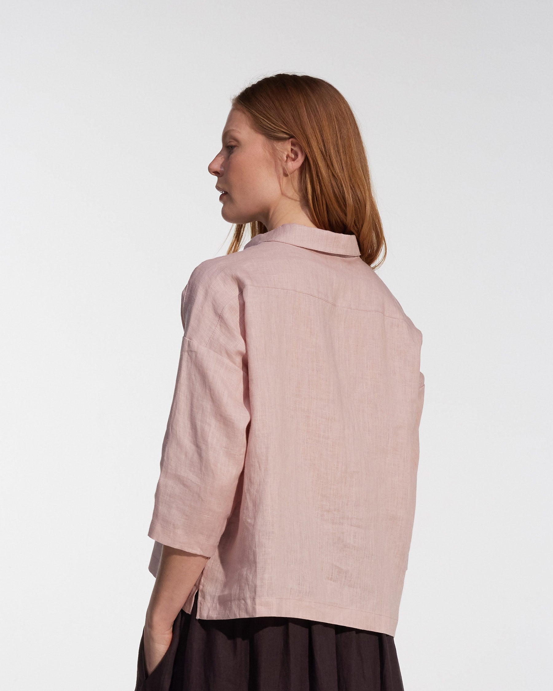 A woman with long light brown hair models the Pyne and Smith Clothiers Boxy Cropped Linen Blouse No.22 in Sepia Rose, showing its oversized fit and loose three-quarter sleeves from the side and back against a white background.