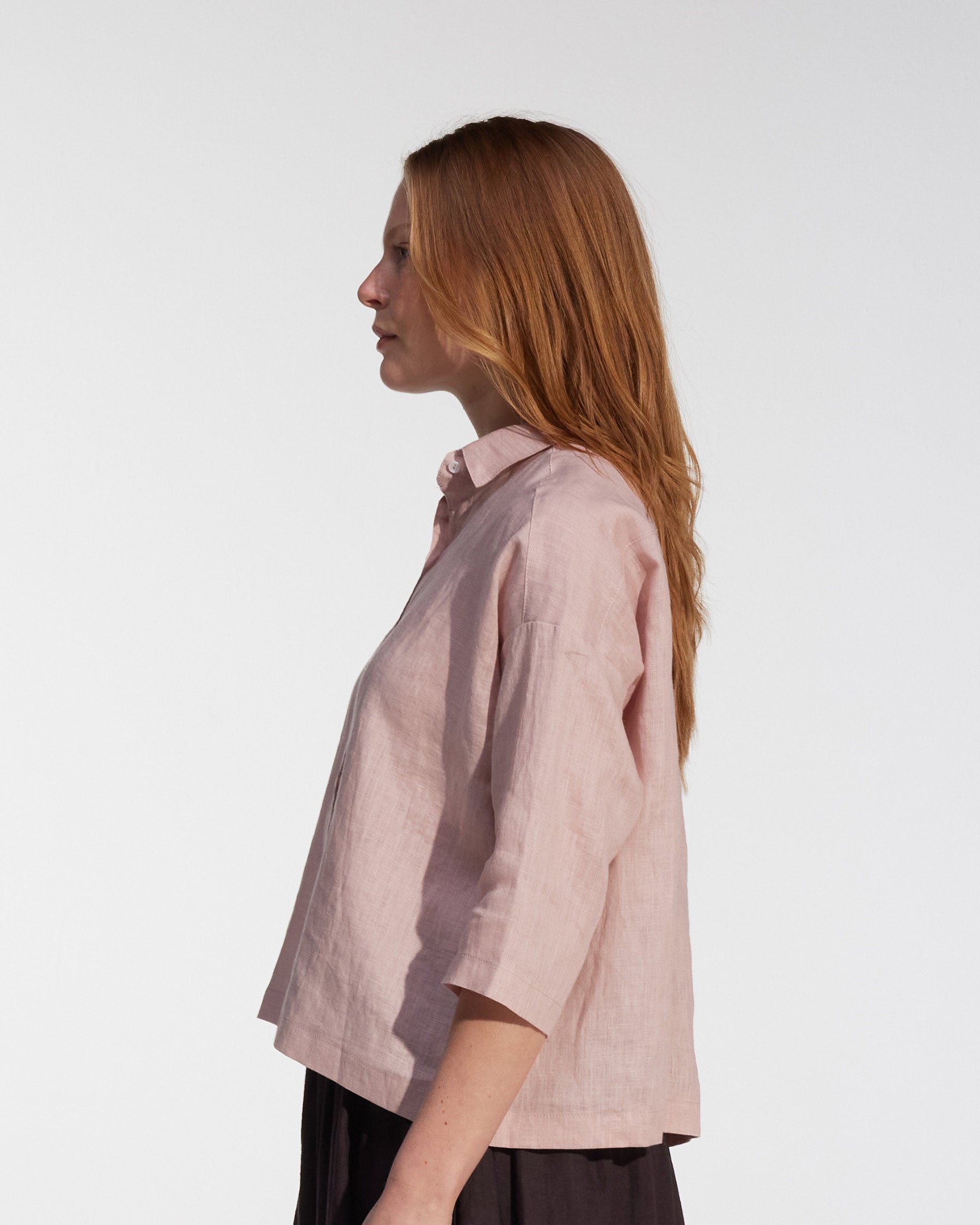 A woman with long, straight, light brown hair stands in profile against a plain white background, wearing the Pyne and Smith Clothiers Boxy Cropped Linen Blouse No.22 in Sepia Rose and a dark skirt.