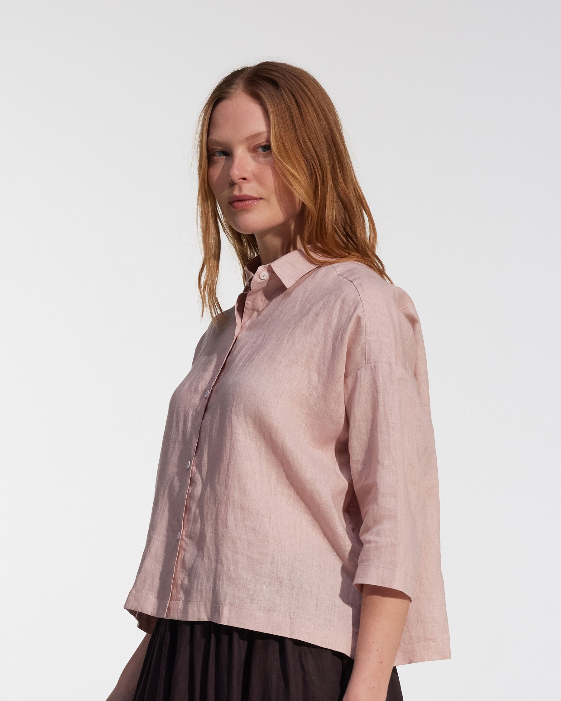 A woman with light brown hair wears the Boxy Cropped Linen Blouse No.22 in Sepia Rose by Pyne and Smith Clothiers, paired with a black skirt, standing against a plain white background.