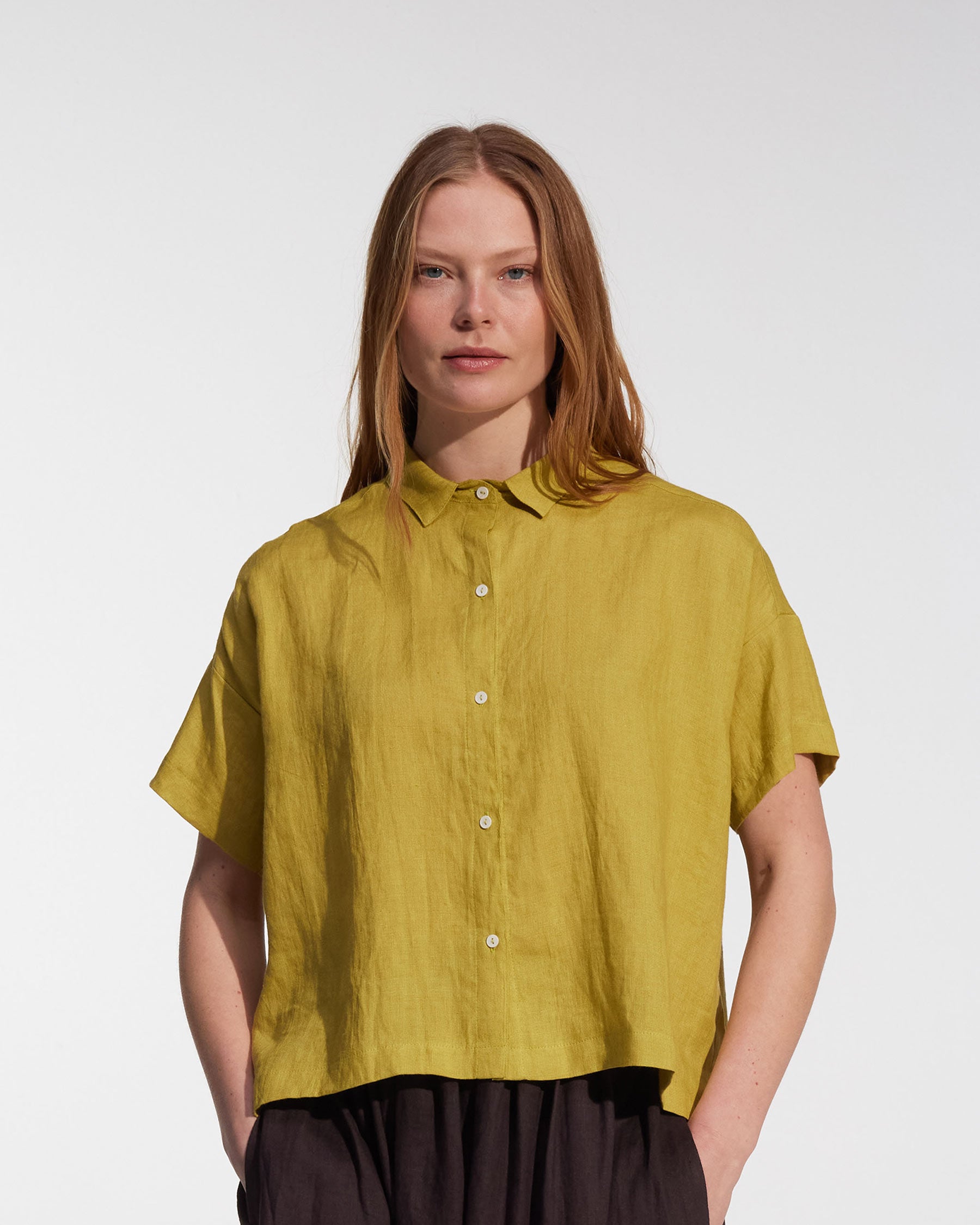 A woman with straight, light brown hair is wearing the Pyne and Smith Clothiers Cropped Linen Blouse No.24 in Apple with dark pants, standing against a plain white background.