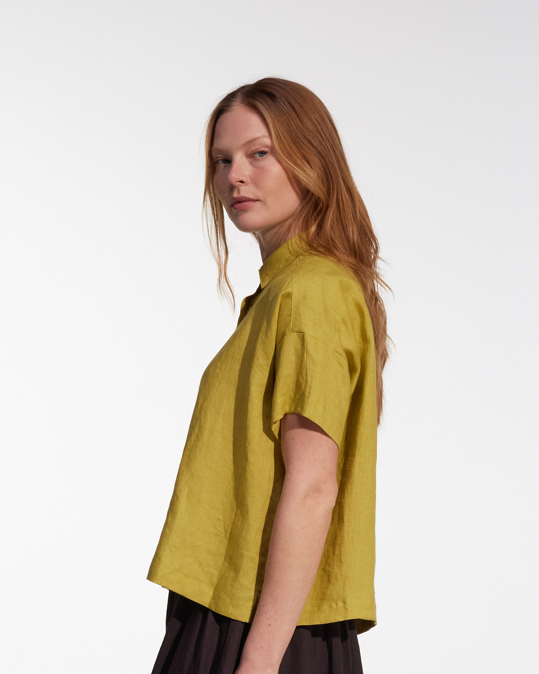A woman with long, straight, light brown hair stands sideways against a plain white background, wearing the Cropped Linen Blouse No.24 in Apple by Pyne and Smith Clothiers and a dark skirt.