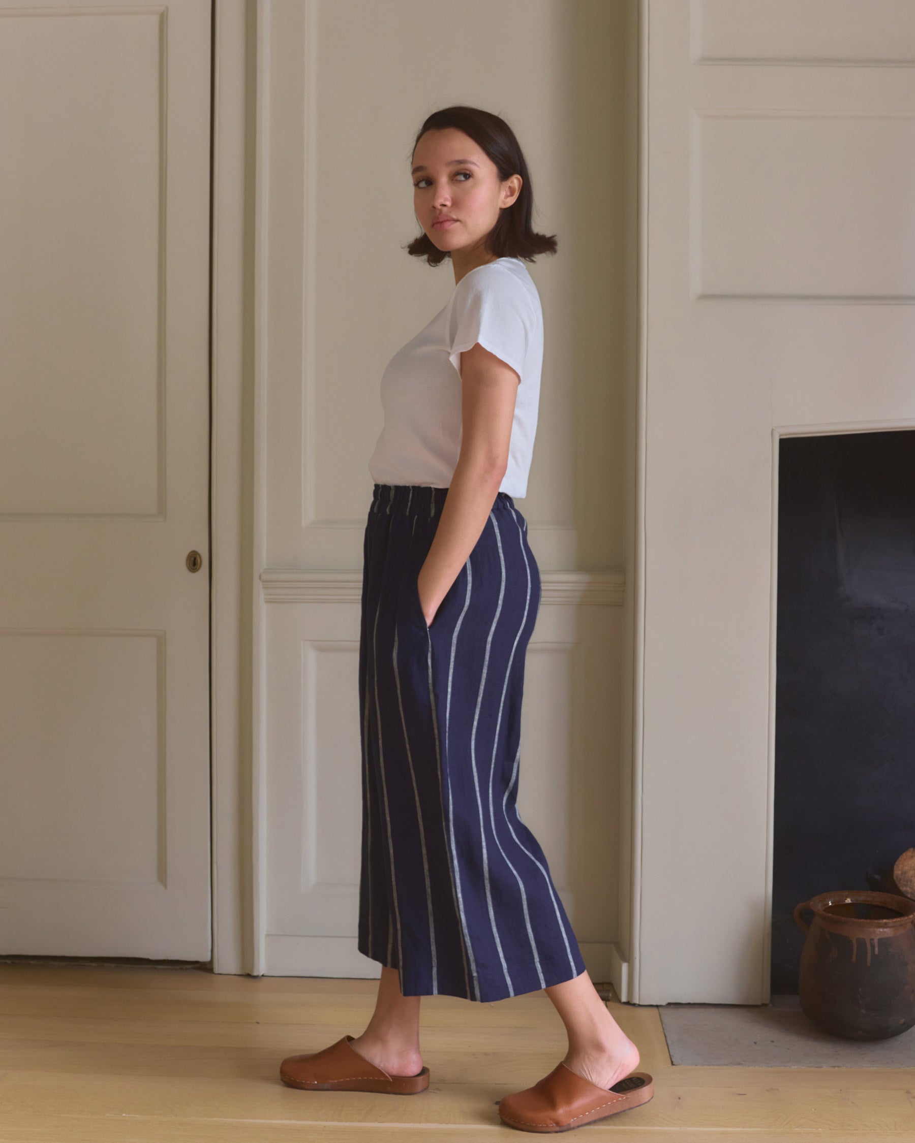 A woman stands indoors in profile, wearing a white t-shirt and Pyne and Smith Clothiers Culottes No.11 Linen Cropped Pants in Blundell Stripe, paired with brown slip-ons. She has her hands in her pockets and looks toward the camera.