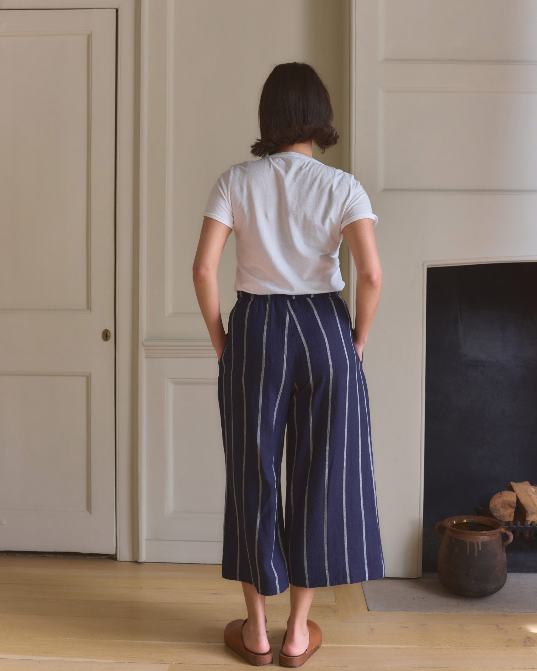 A person with short brown hair stands indoors facing away, wearing a white t-shirt and Pyne and Smith Clothiers Culottes No.11 Linen Cropped Pants in Blundell Stripe, hands in their pockets by a fireplace.