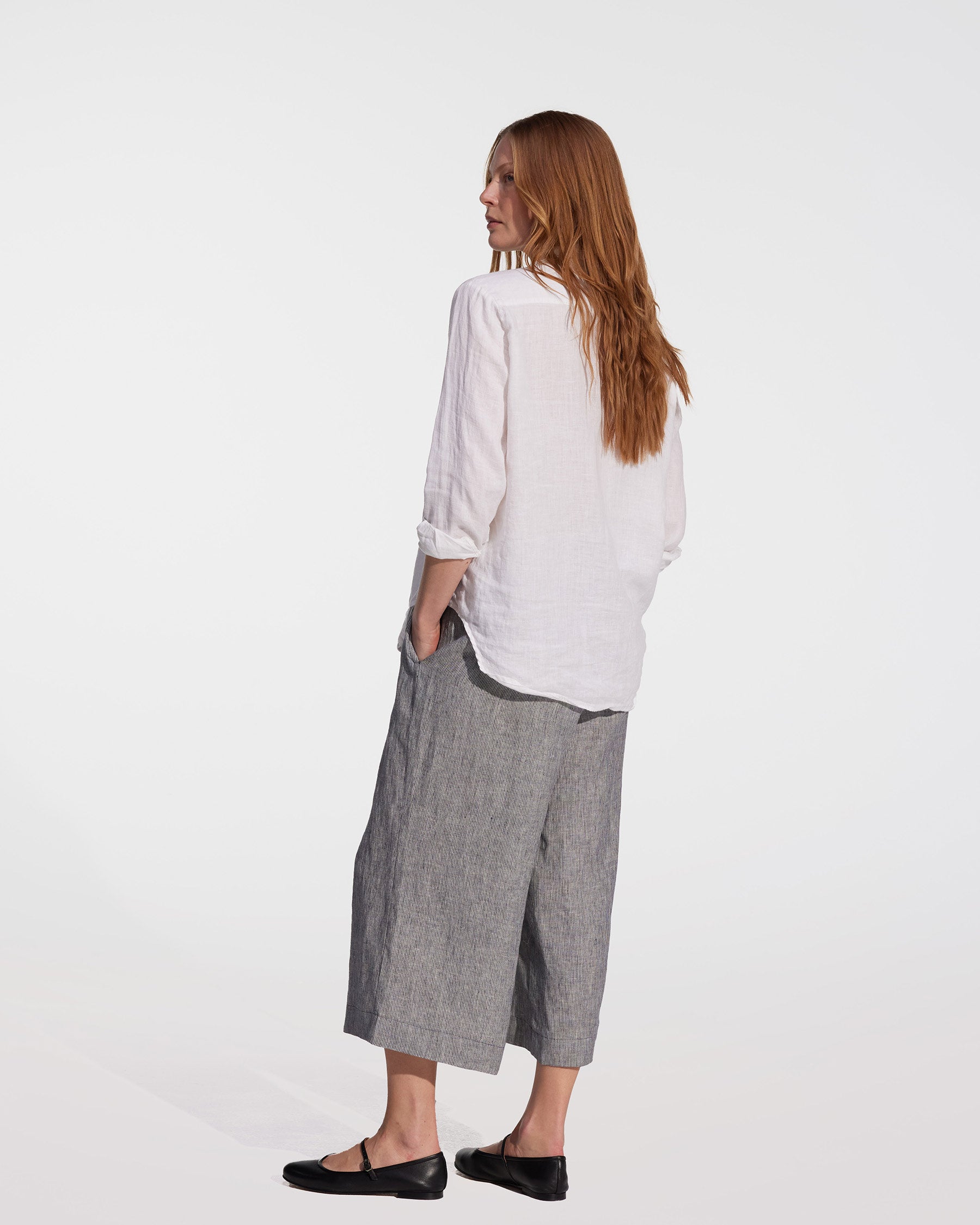 A woman with long red hair stands sideways, looking away. She wears a loose white shirt, black flats, and Linen Culottes No.11 in Cooke's Stripe by Pyne and Smith Clothiers against a plain white background.