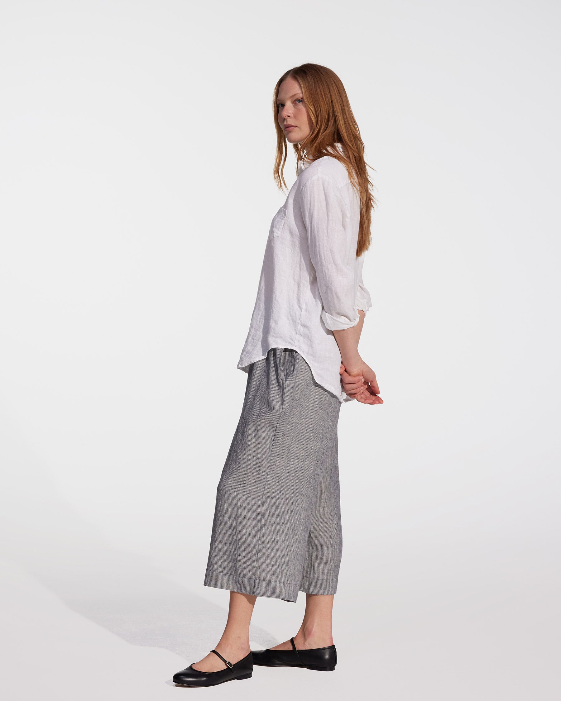 A woman with long red hair, wearing a white blouse and Pyne and Smith Clothiers' Linen Culottes No.11 in Cooke's Stripe, stands sideways against a plain white background with her hands clasped behind her back.