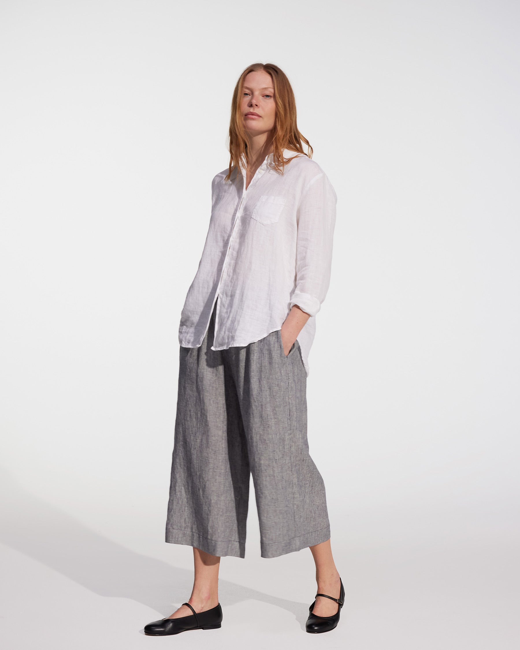A woman stands against a white background wearing a loose white button-up shirt and Pyne and Smith Clothiers' Linen Culottes No.11 in Cooke's Stripe, paired with black Mary Jane flats, her hands in her pockets and expression neutral.