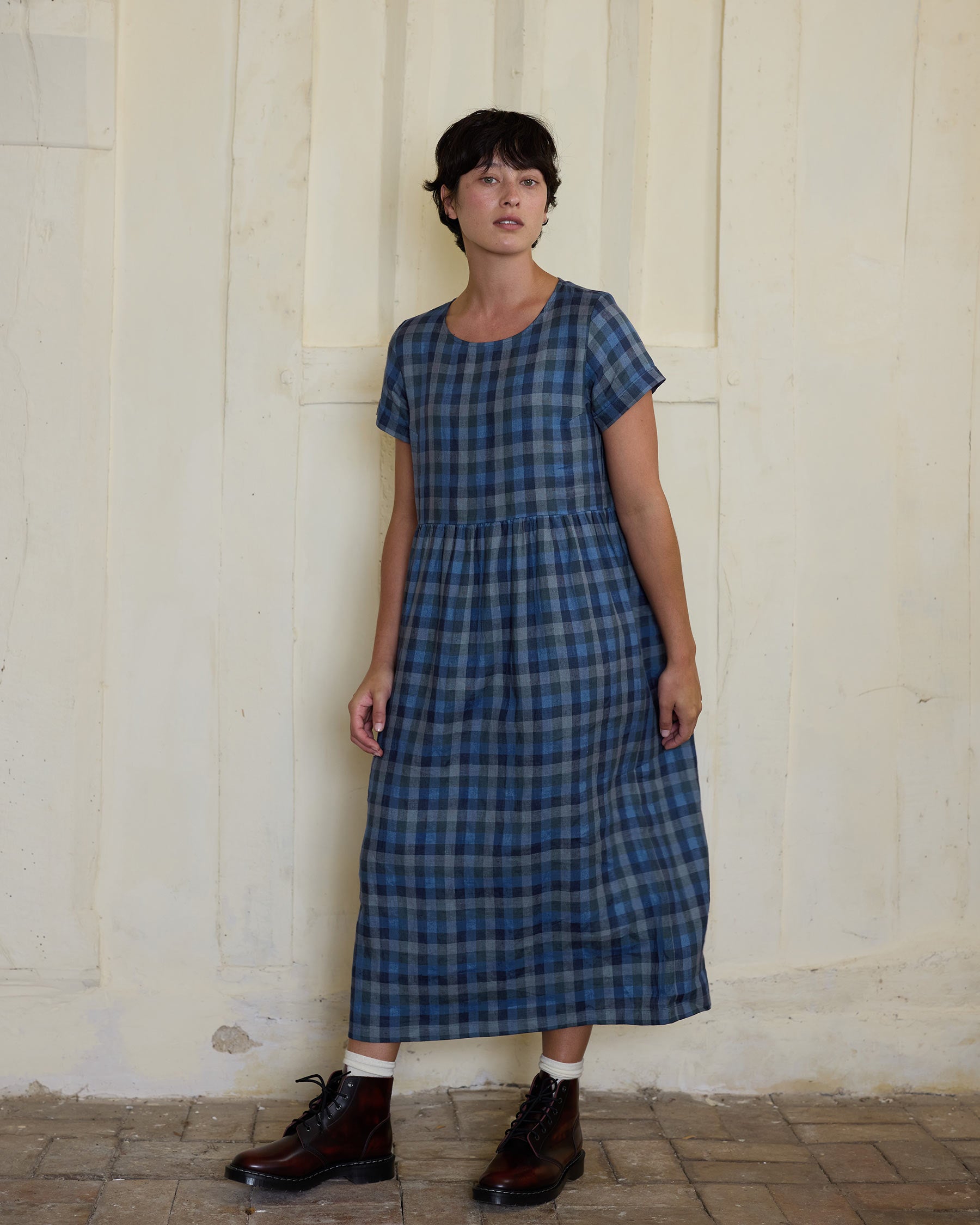 A person with short dark hair wears the Pyne and Smith Clothiers No.17 Full Length Linen Tea Dress in Blue Melange and brown lace-up boots, standing indoors against a pale, textured wall with vertical indentations.