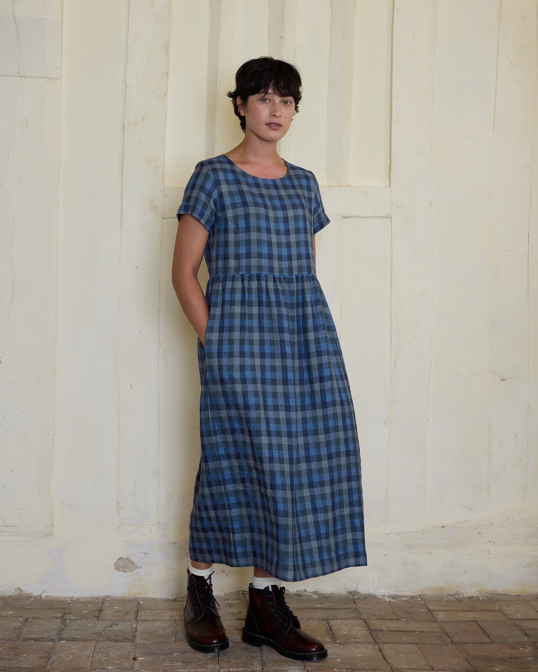 A person with short dark hair stands indoors against a light wall, wearing the Pyne and Smith Clothiers No.17 Full Length Linen Tea Dress in Blue Melange and brown lace-up boots, hands in pockets, looking at the camera.