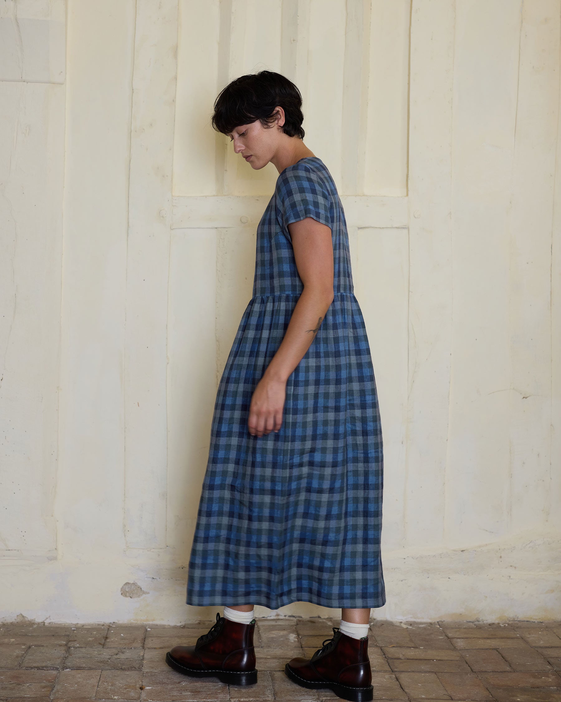 A person with short dark hair wears the Pyne and Smith Clothiers No.17 Full Length Linen Tea Dress in Blue Melange with brown boots, standing in profile against a textured cream wall and tiled floor.