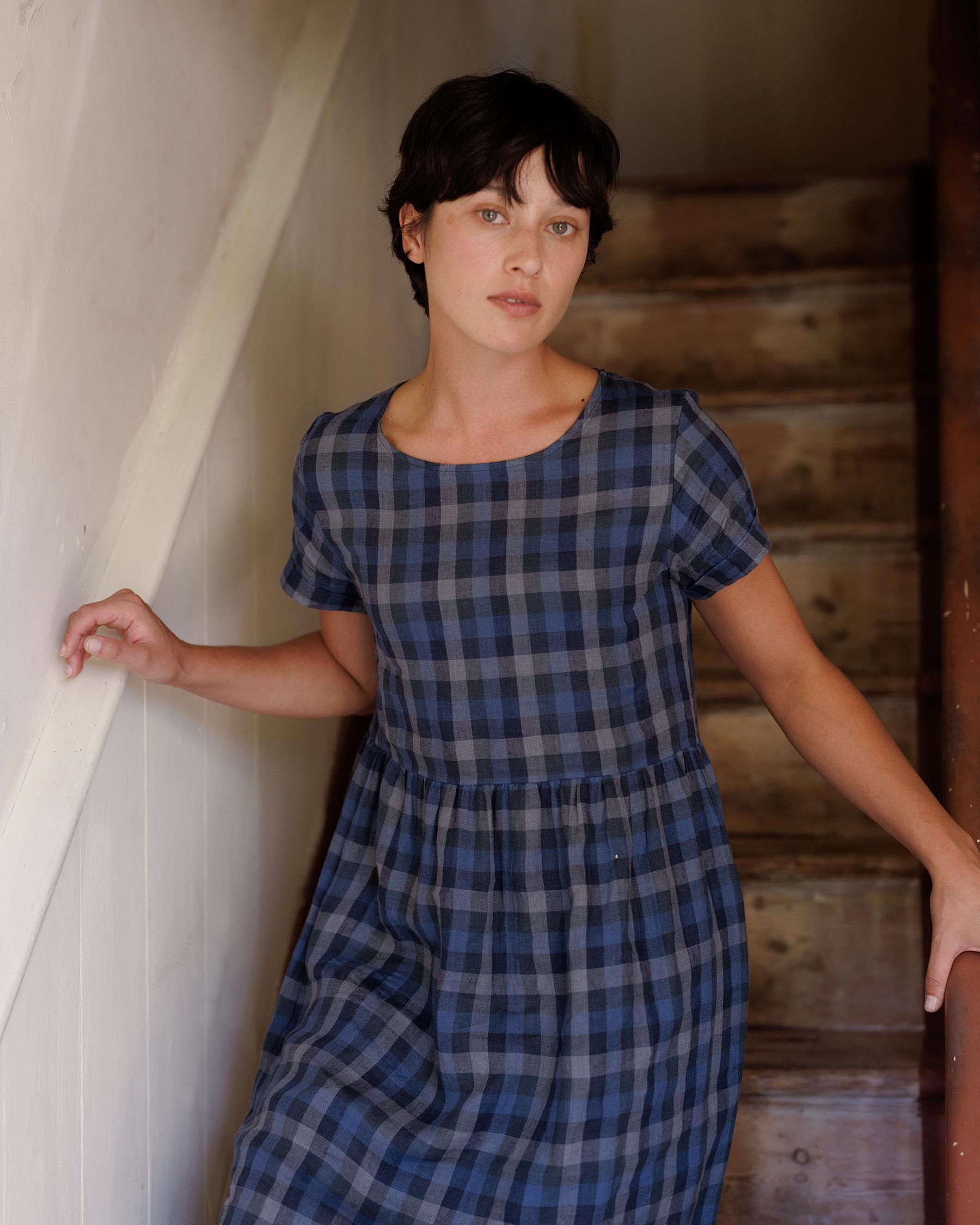 A woman with short dark hair wears the Pyne and Smith Clothiers No.17 Full Length Linen Tea Dress in Blue Melange, standing on wooden stairs, holding the railing with one hand and touching the wall, calmly facing the camera.