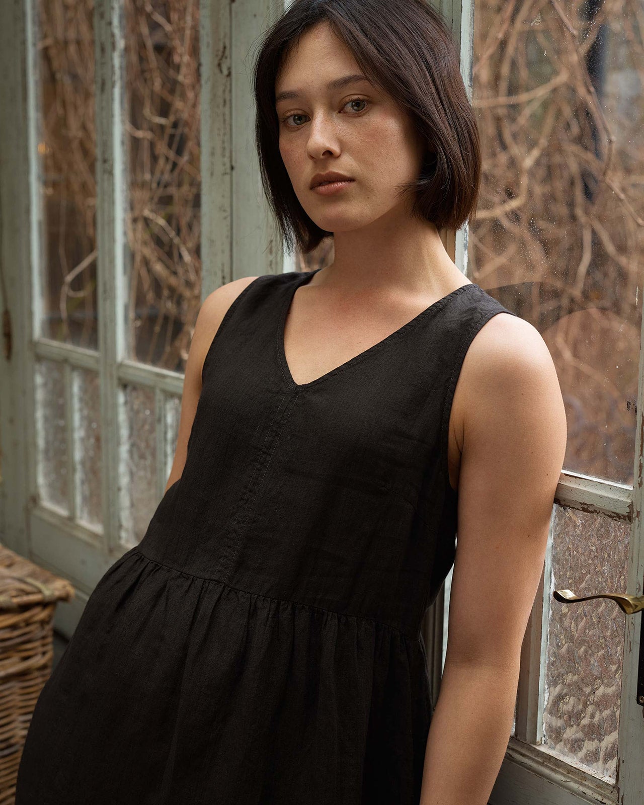 A woman with short dark hair stands indoors by a window, wearing the Pyne and Smith Clothiers Size Medium #676 No.21 V Neck Sleeveless Linen Dress in Black. She looks at the camera with a neutral expression; leafless branches are visible outside.