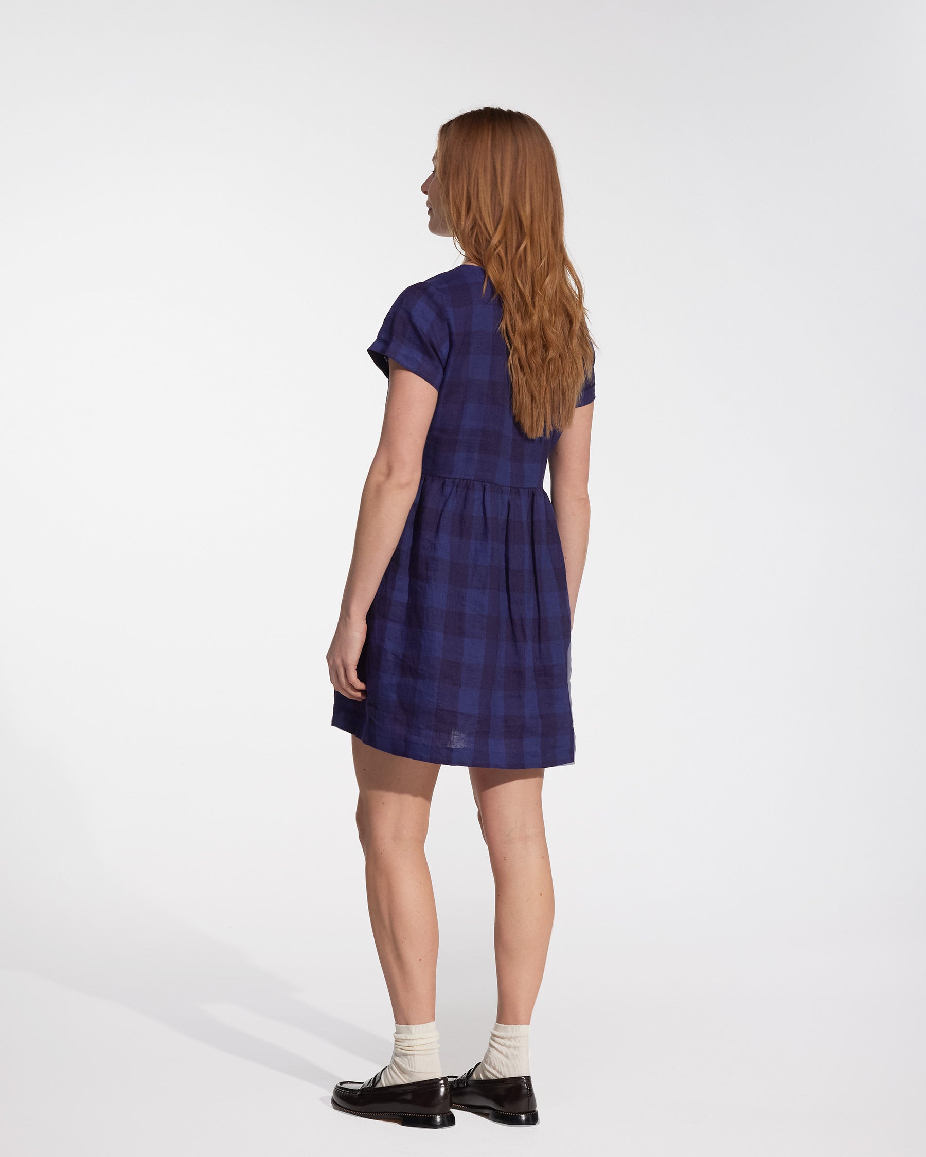 A woman with long red hair faces away, wearing the Pyne and Smith Clothiers No.10 Button Up Linen Dress in Midsommar Night Check, styled with white socks and black shoes, set against a plain white background.