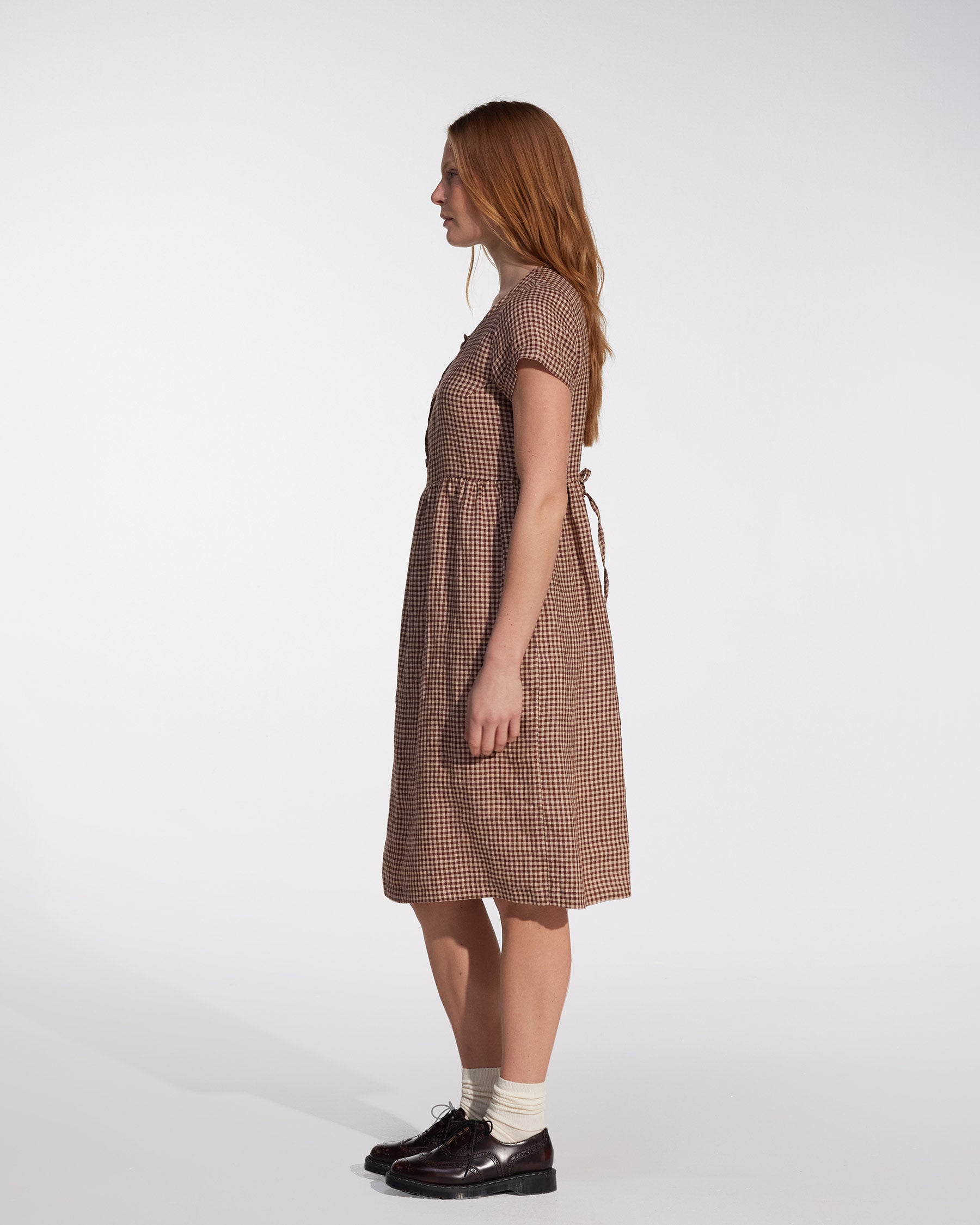 A woman with long hair stands in profile wearing the Pyne and Smith Clothiers No.11 Classic Button Up Linen Dress in Topsham Check, styled with cream socks and dark shoes, against a plain white background.