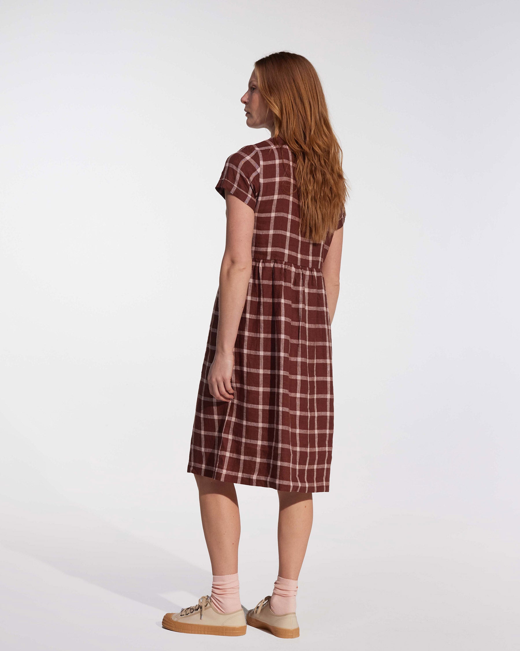 A woman with long red hair, facing away, wears the Pyne and Smith Clothiers No.15 Linen Day Dress in Neapolitan with side pockets, paired with beige sneakers and light pink socks against a plain white background.
