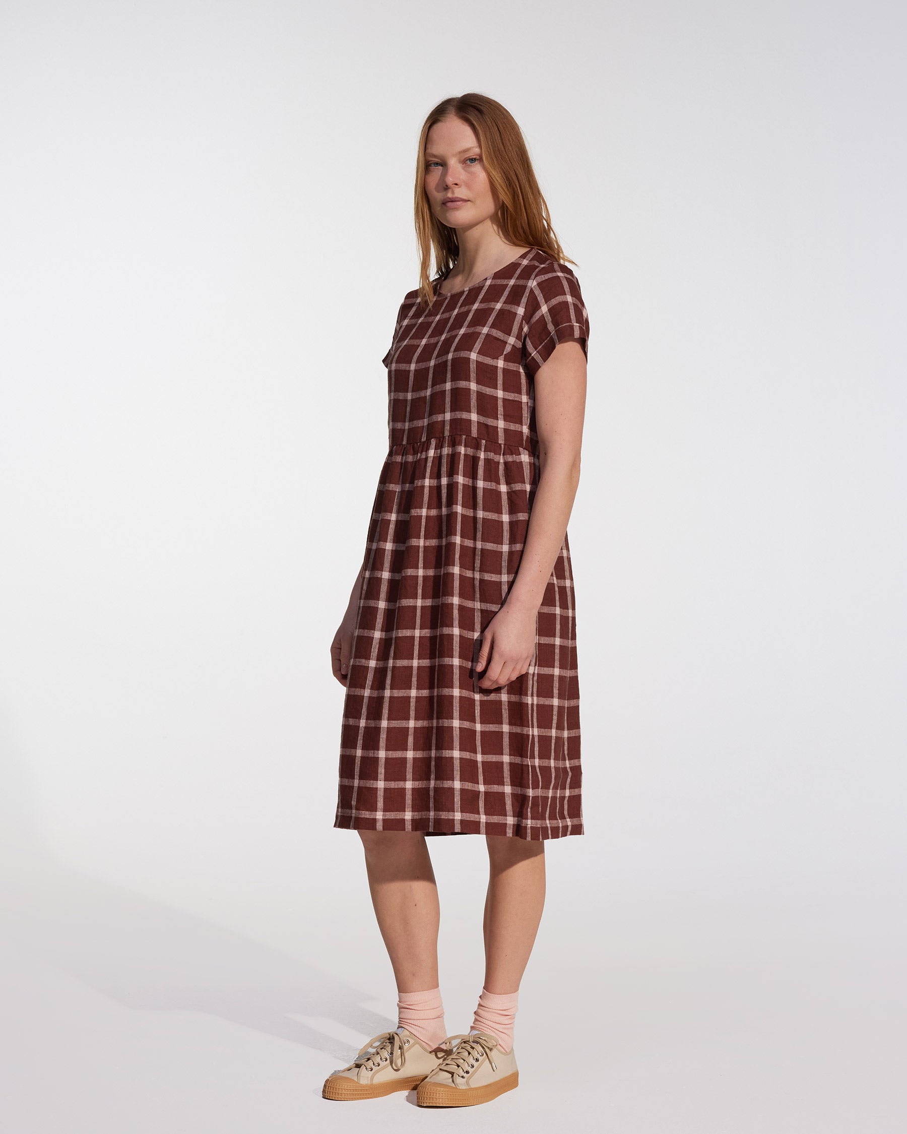 A woman with straight, shoulder-length hair and a neutral expression stands against a white background wearing the Pyne and Smith Clothiers No.15 Linen Day Dress in Neapolitan, with side pockets, light pink socks, and beige sneakers.