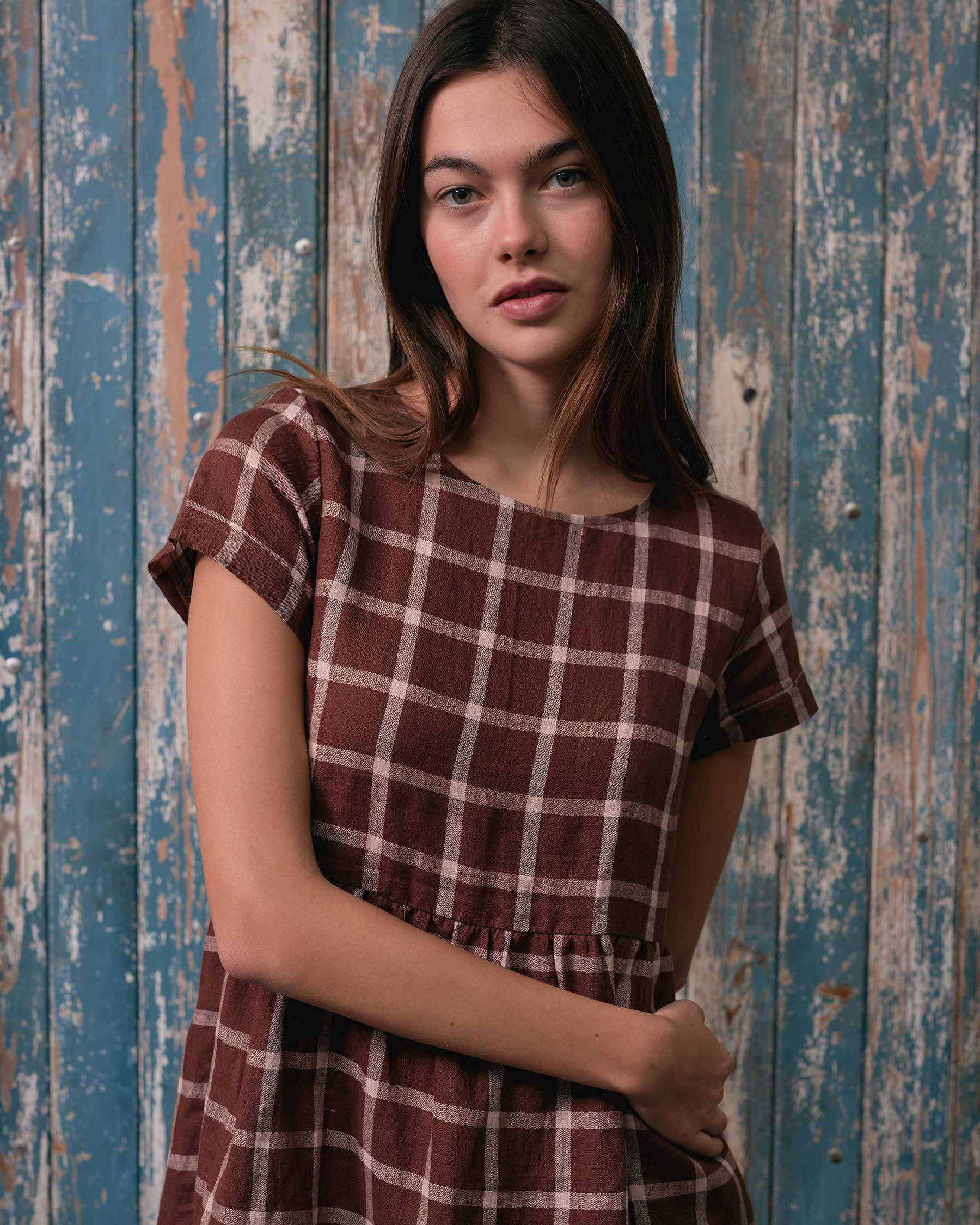 A woman with long brown hair wears the Pyne and Smith Clothiers No.15 Linen Day Dress in Neapolitan, a short-sleeved brown and white plaid midi with side pockets, standing in front of a weathered blue wooden wall.