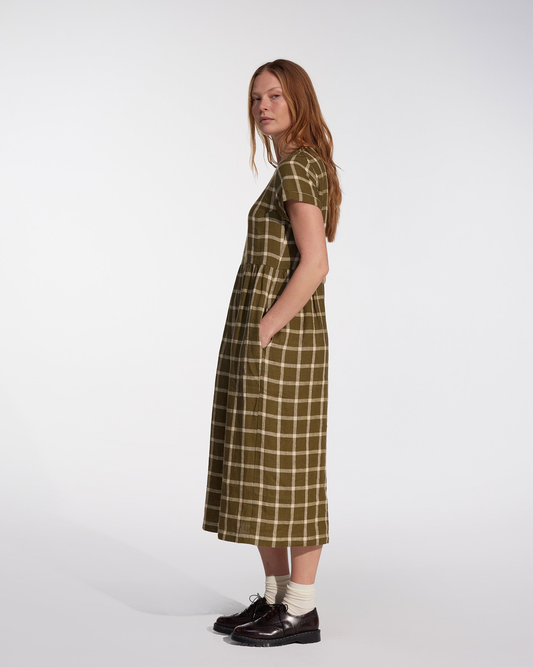 A woman with long red hair stands sideways in the Pyne and Smith Clothiers No.17 Full Length Linen Tea Dress in Celtic Plaid, paired with white socks and black shoes, set against a plain white background.