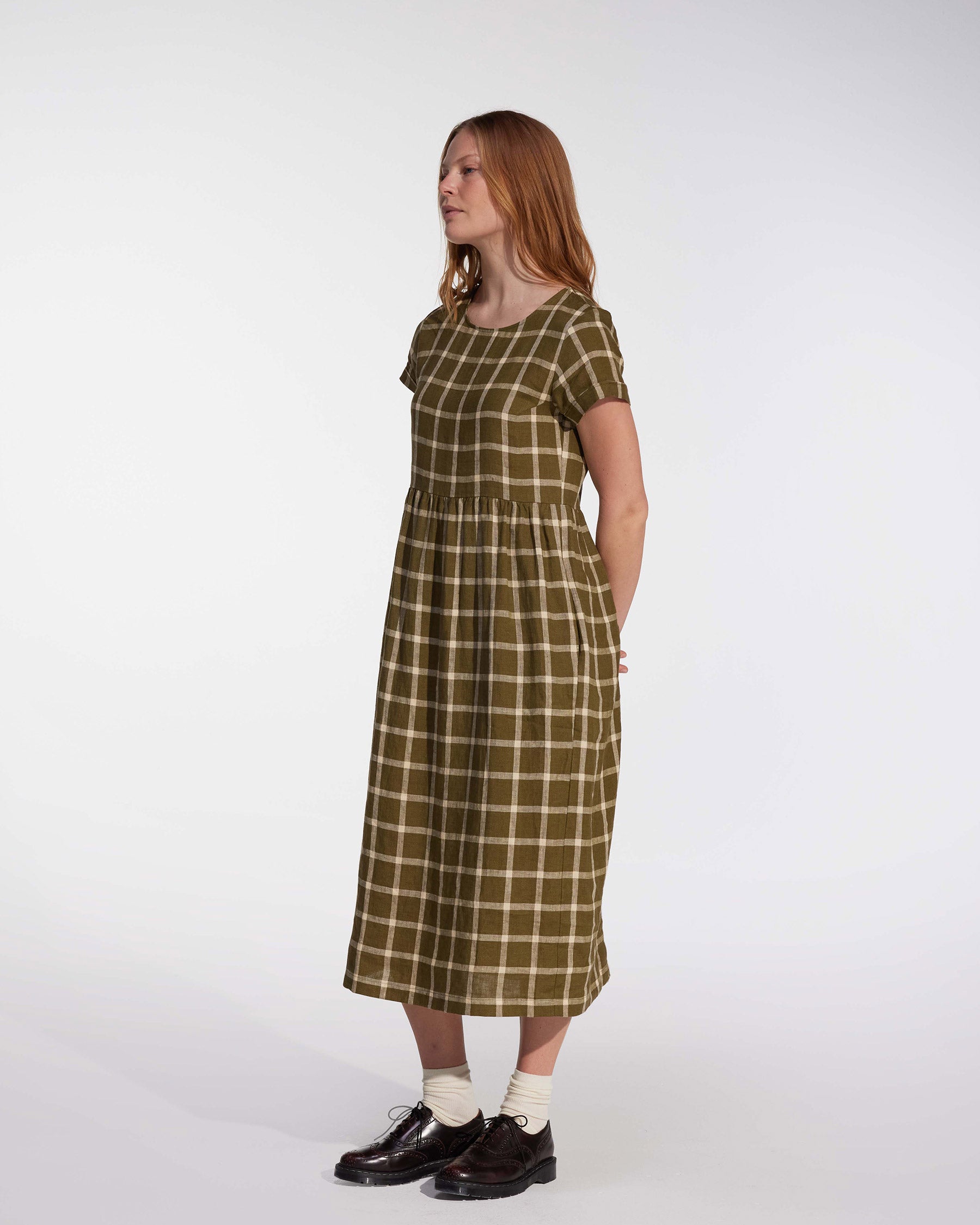 A woman with long red hair stands sideways in the No.17 Full Length Linen Tea Dress in Celtic Plaid by Pyne and Smith Clothiers, paired with white socks and dark brown shoes, against a plain light background.