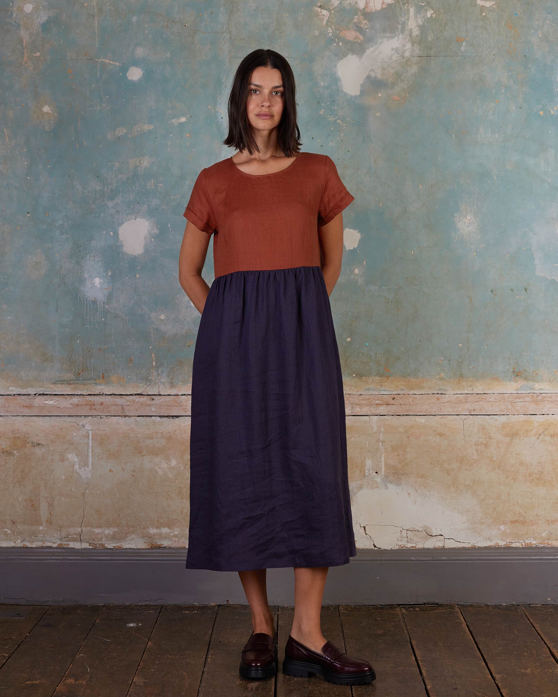 A woman stands indoors against a weathered blue and beige wall, wearing the No.17 Full Length Linen Tea Dress in Nicchio by Pyne and Smith Clothiers, paired with brown loafers, looking forward with her hands behind her back.
