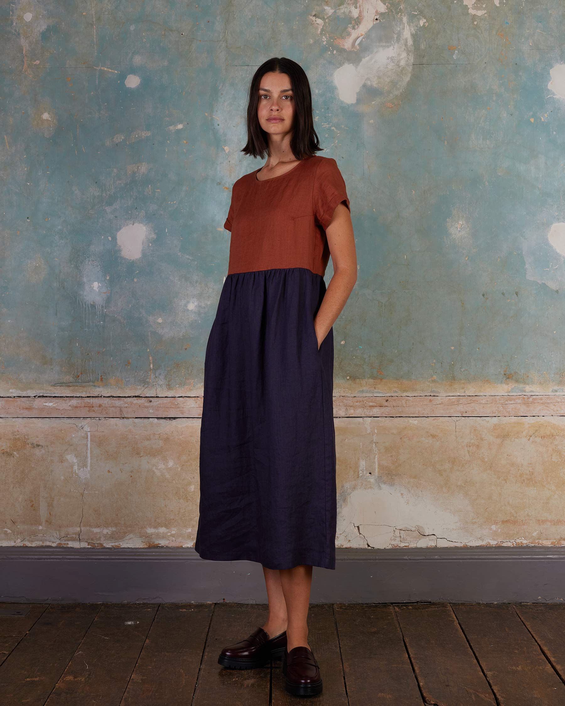 A woman stands indoors against a textured wall, wearing Pyne and Smith Clothiers' No.17 Full Length Linen Tea Dress in Nicchio, styled with a short-sleeved rust top and black shoes, looking ahead with a neutral expression.
