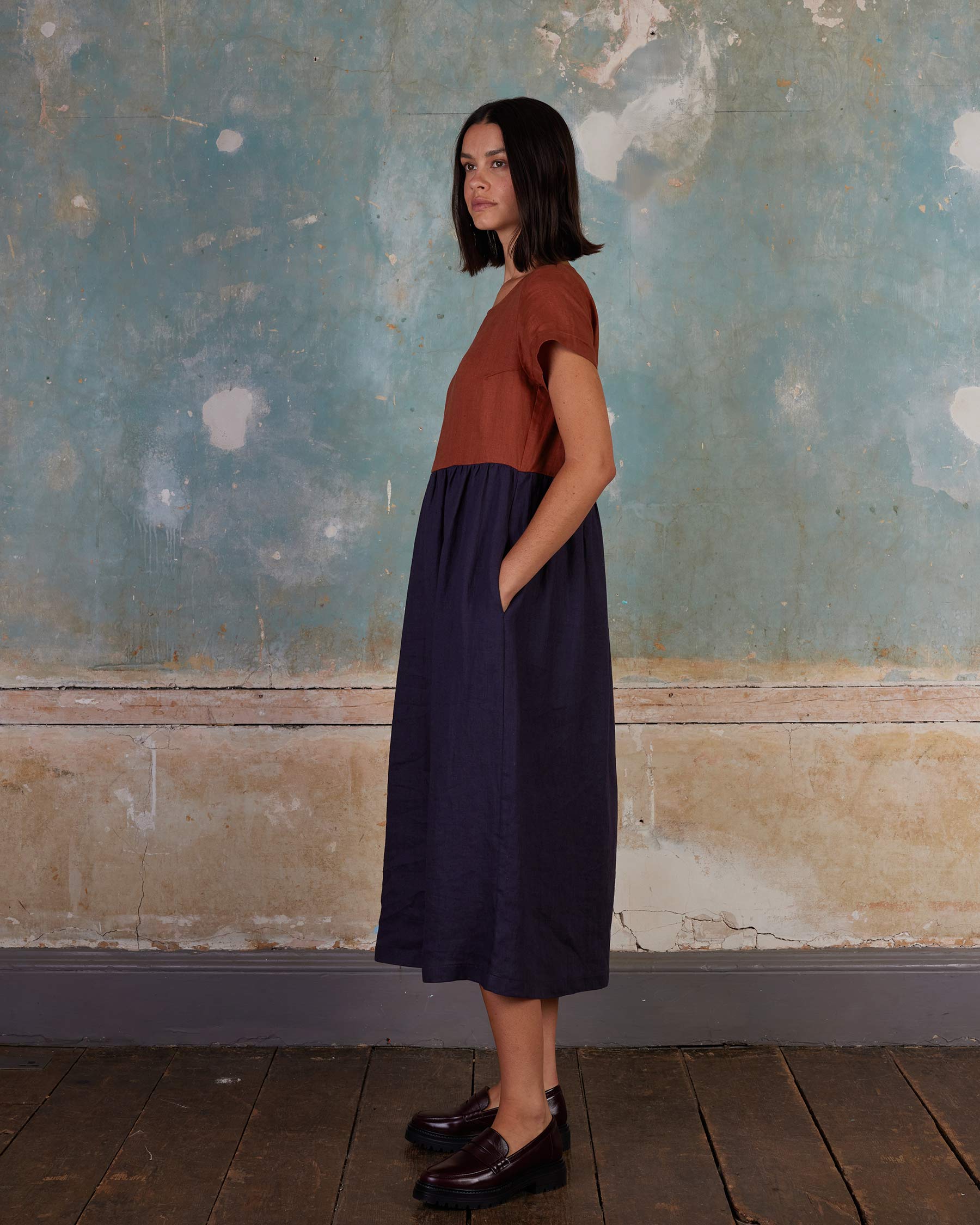 A woman with shoulder-length dark hair stands in profile against a textured wall, wearing the Pyne and Smith Clothiers No.17 Full Length Linen Tea Dress in Nicchio and dark loafers on a wooden floor.