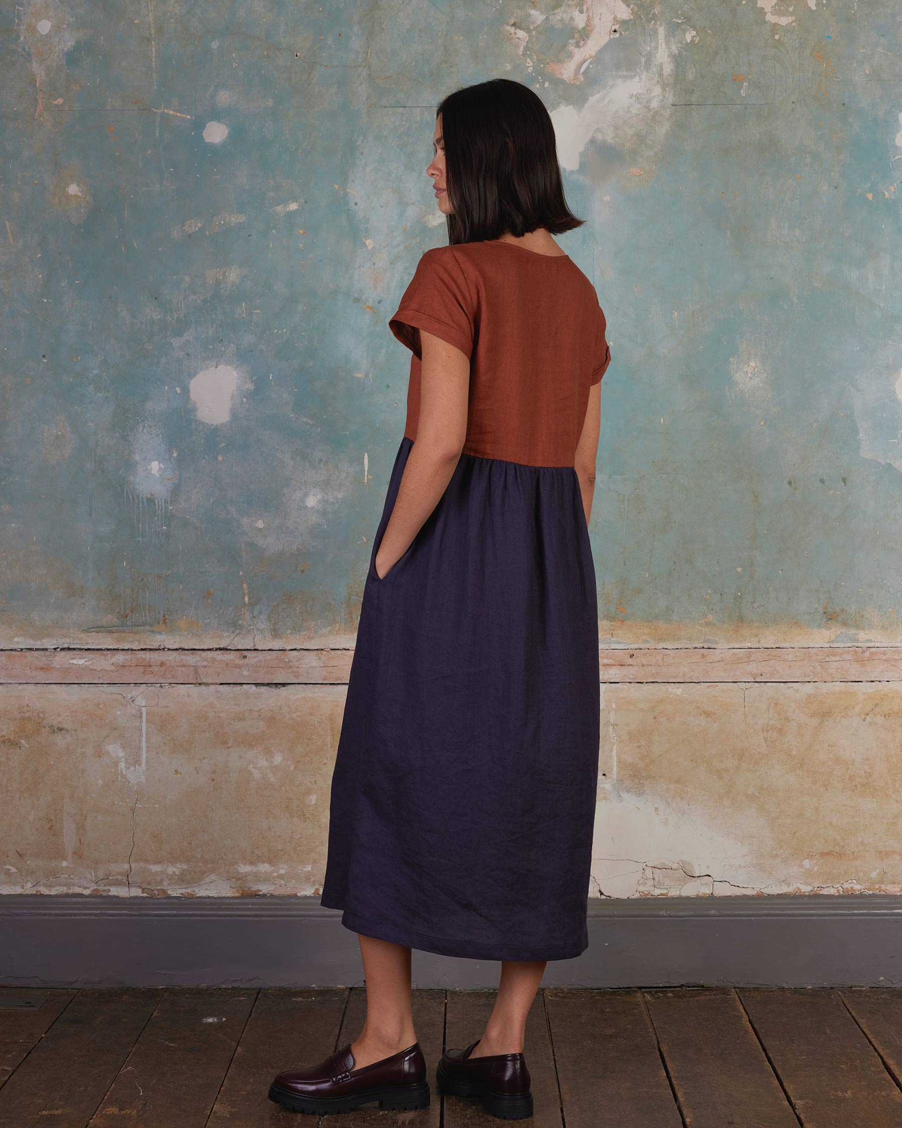 A woman with straight dark hair stands sideways by a blue and beige wall, wearing the No.17 Full Length Linen Tea Dress in Nicchio from Pyne and Smith Clothiers, paired with brown loafers and hands tucked casually in her pockets.