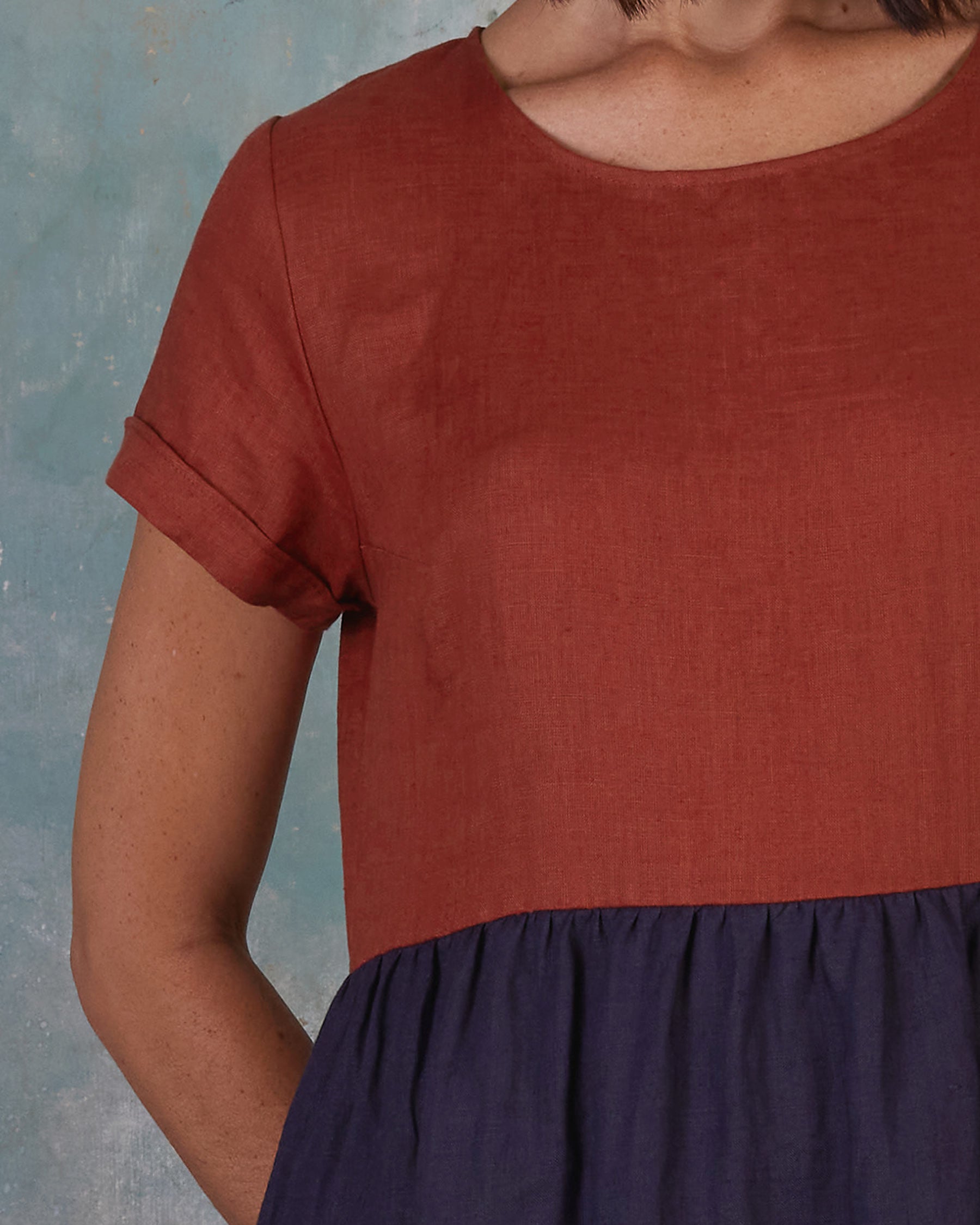 A person in the No.17 Full Length Linen Tea Dress in Nicchio by Pyne and Smith Clothiers, featuring a rust top and navy skirt, stands before a blue textured background with only part of their face and upper body visible.