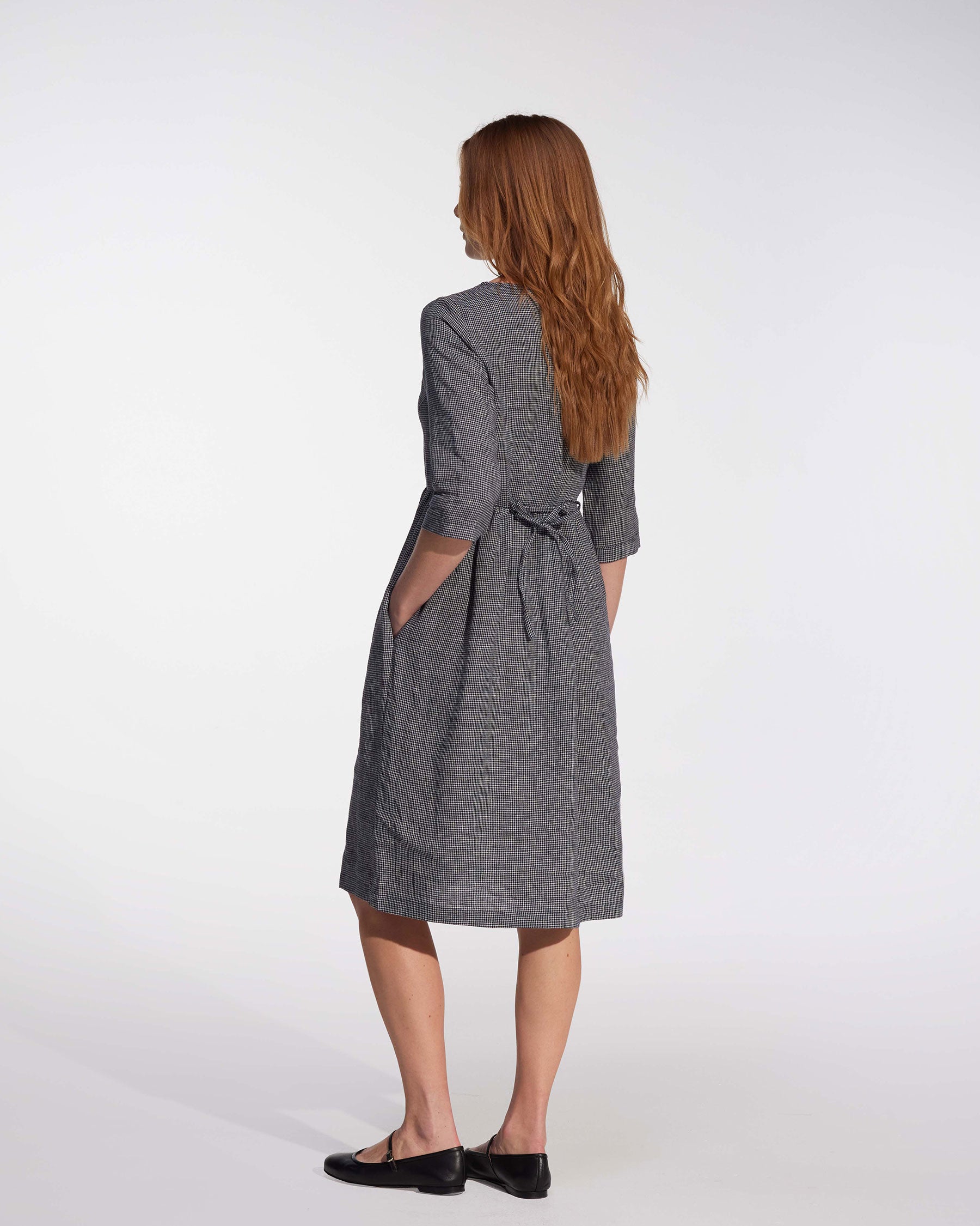 A woman with long, reddish-brown hair faces away, wearing Pyne and Smith Clothiers' No.19 Midi Linen Dress in Brasserie Check and black flats against a plain white background.