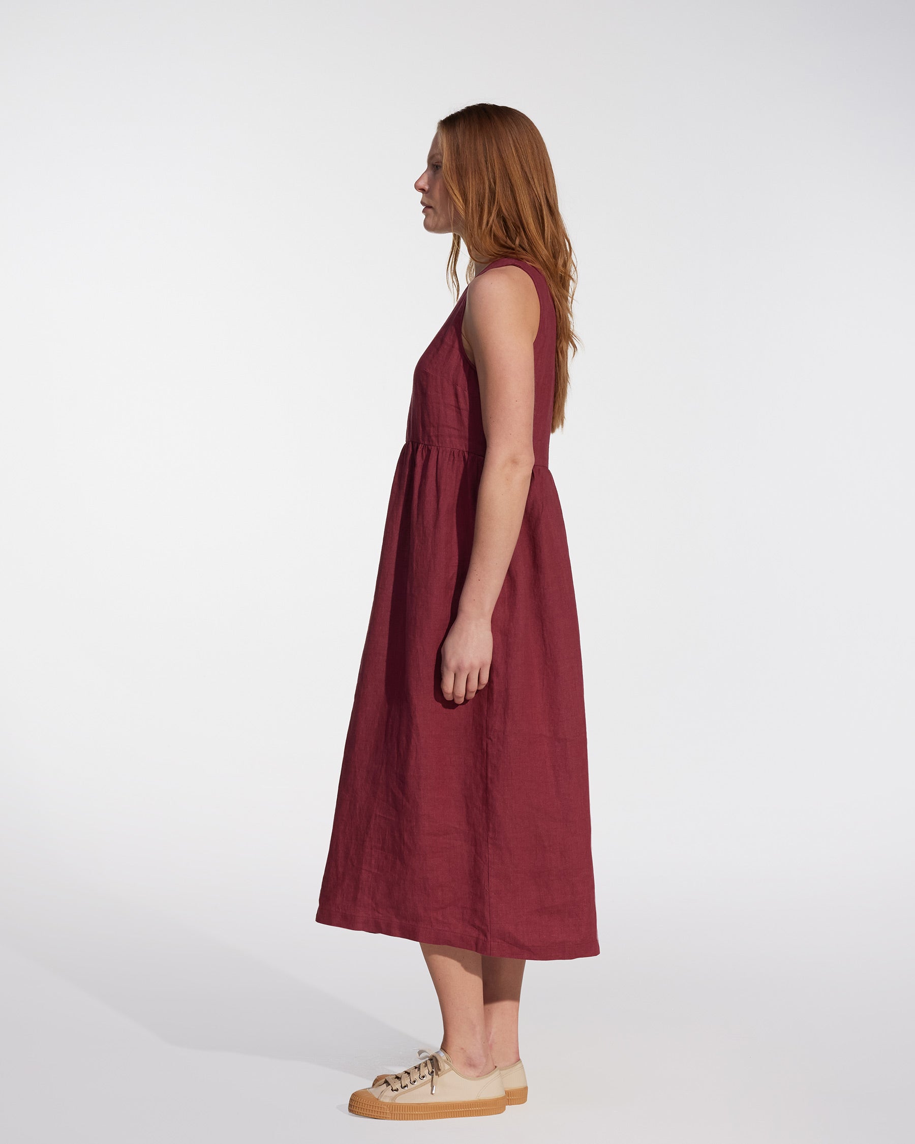 A woman with long red hair stands in profile against a white background, wearing the Pyne and Smith Clothiers No.21 V Neck Sleeveless Linen Dress in Roan and beige sneakers.