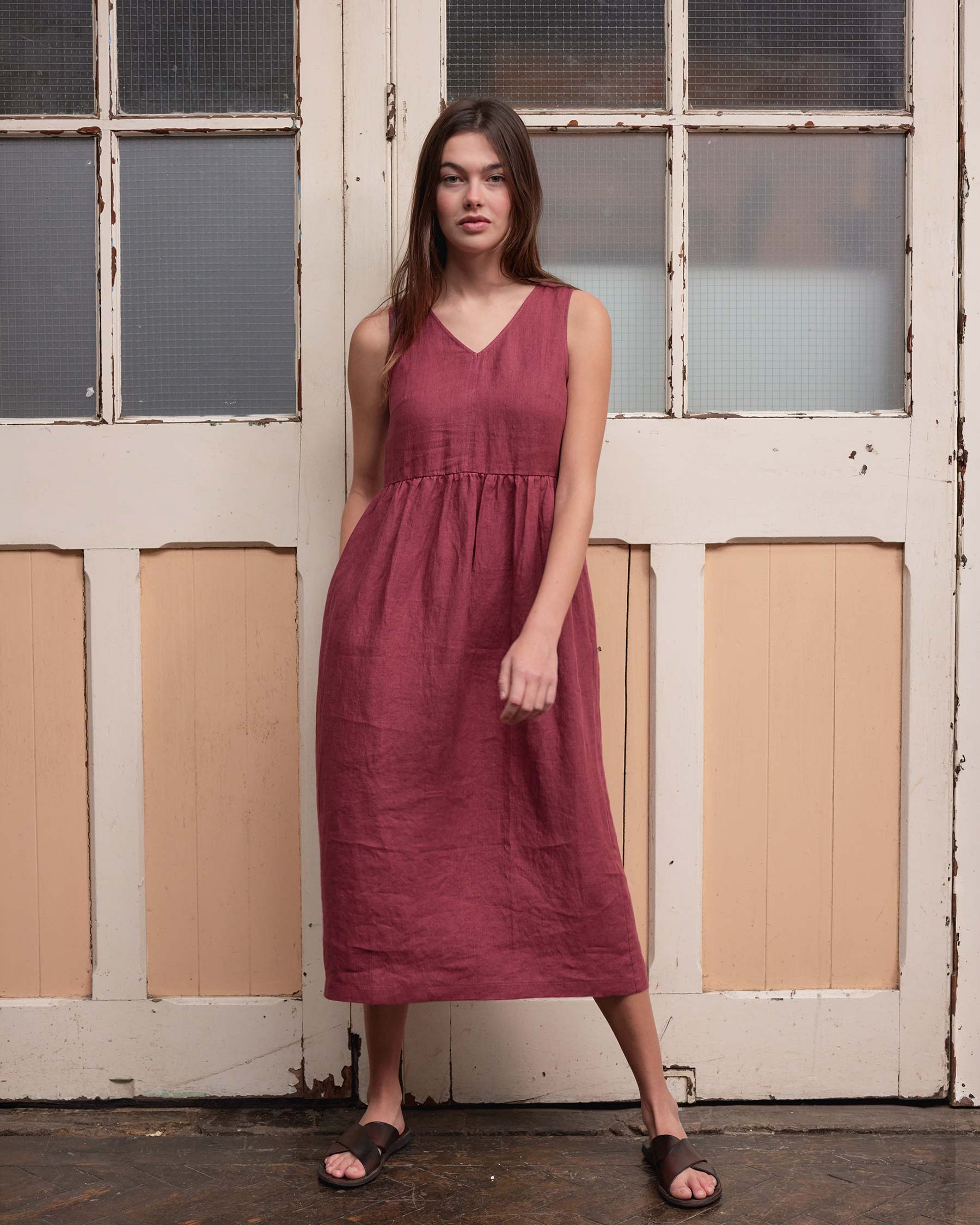 A woman with long brown hair and a relaxed expression stands indoors by old wooden doors, wearing the Pyne and Smith Clothiers No.21 V Neck Sleeveless Linen Dress in Roan with dark sandals.