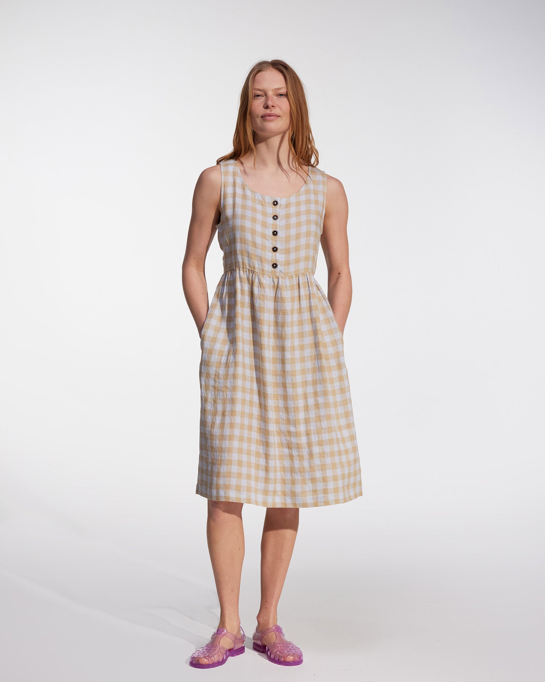 A woman with long hair wears the Pyne and Smith Clothiers No.24 Sleeveless, button-up Linen Dress in St. Agnes Check, featuring front buttons and pockets, paired with purple jelly sandals against a plain white background.