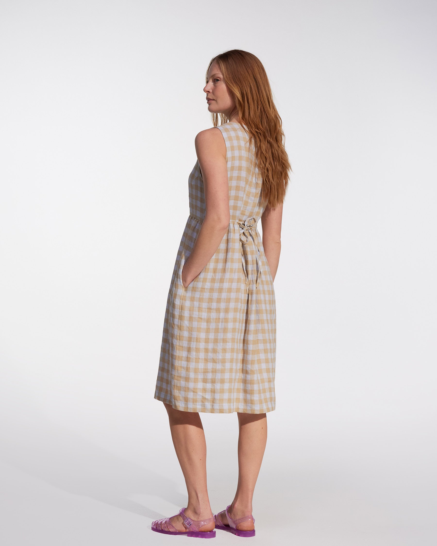 A woman with long red hair poses sideways against a white background, hands in pockets, wearing the Pyne and Smith Clothiers No.24 sleeveless button-up linen dress in St. Agnes Check, styled with clear purple sandals.