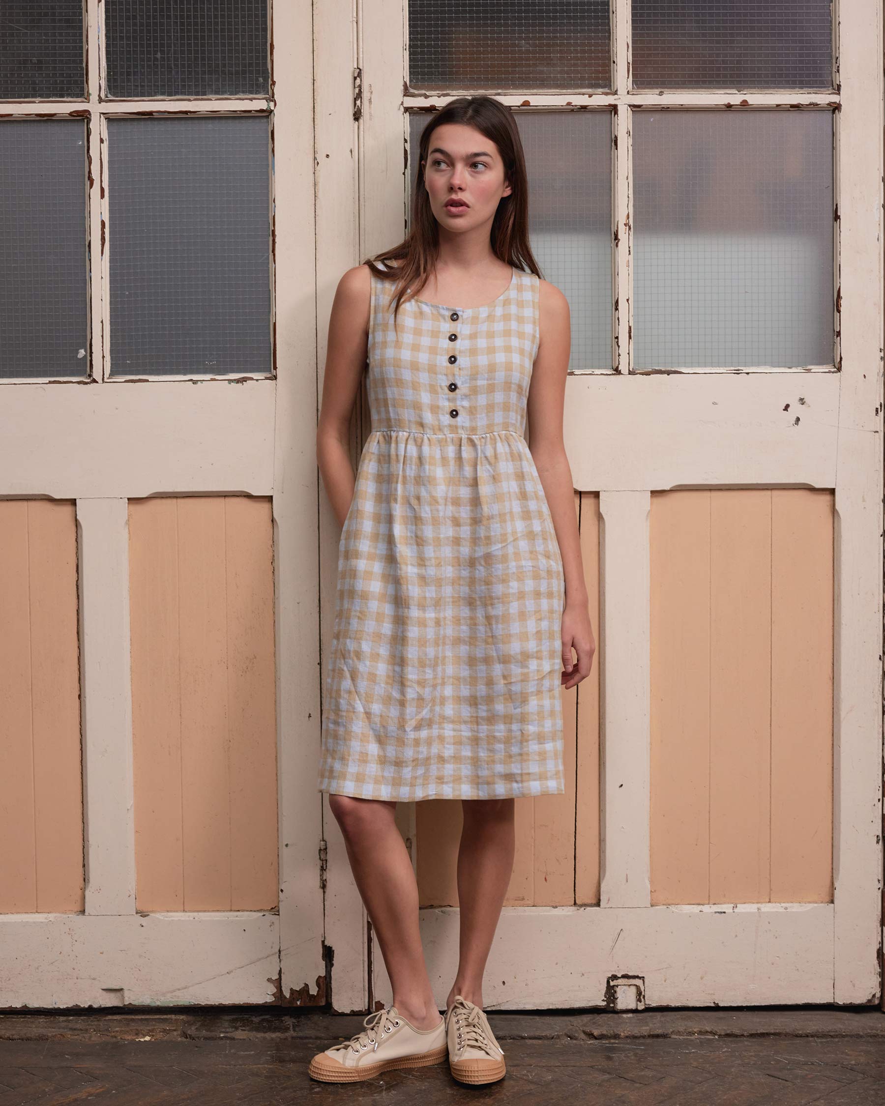 A woman stands before cream and glass-paneled doors, wearing the No.24 Sleeveless, button-up Linen Dress in St. Agnes Check by Pyne and Smith Clothiers with beige sneakers.