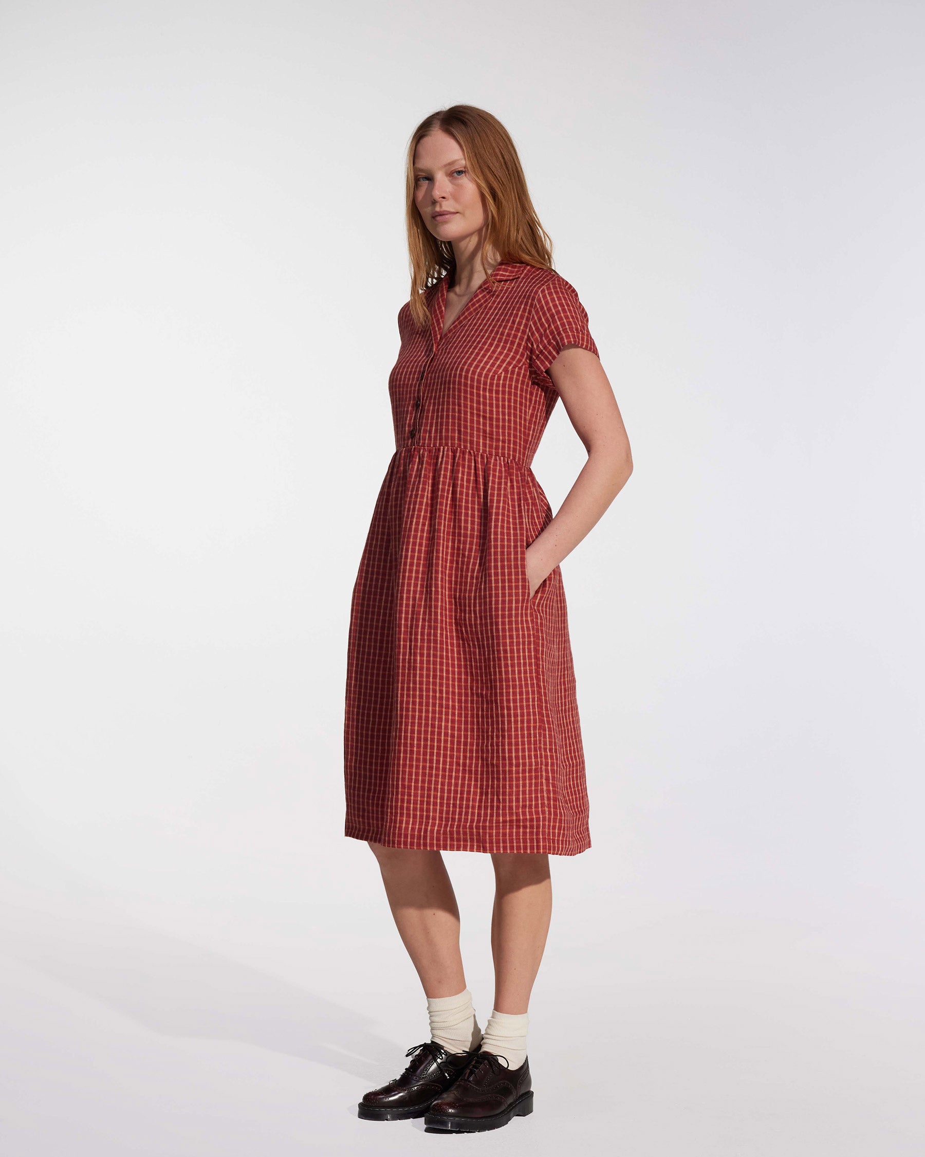 A woman with long reddish hair wears the Pyne and Smith Clothiers No.30 Collared Linen Dress in Pomodoro Plaid, paired with white socks and black shoes, standing against a plain light background.