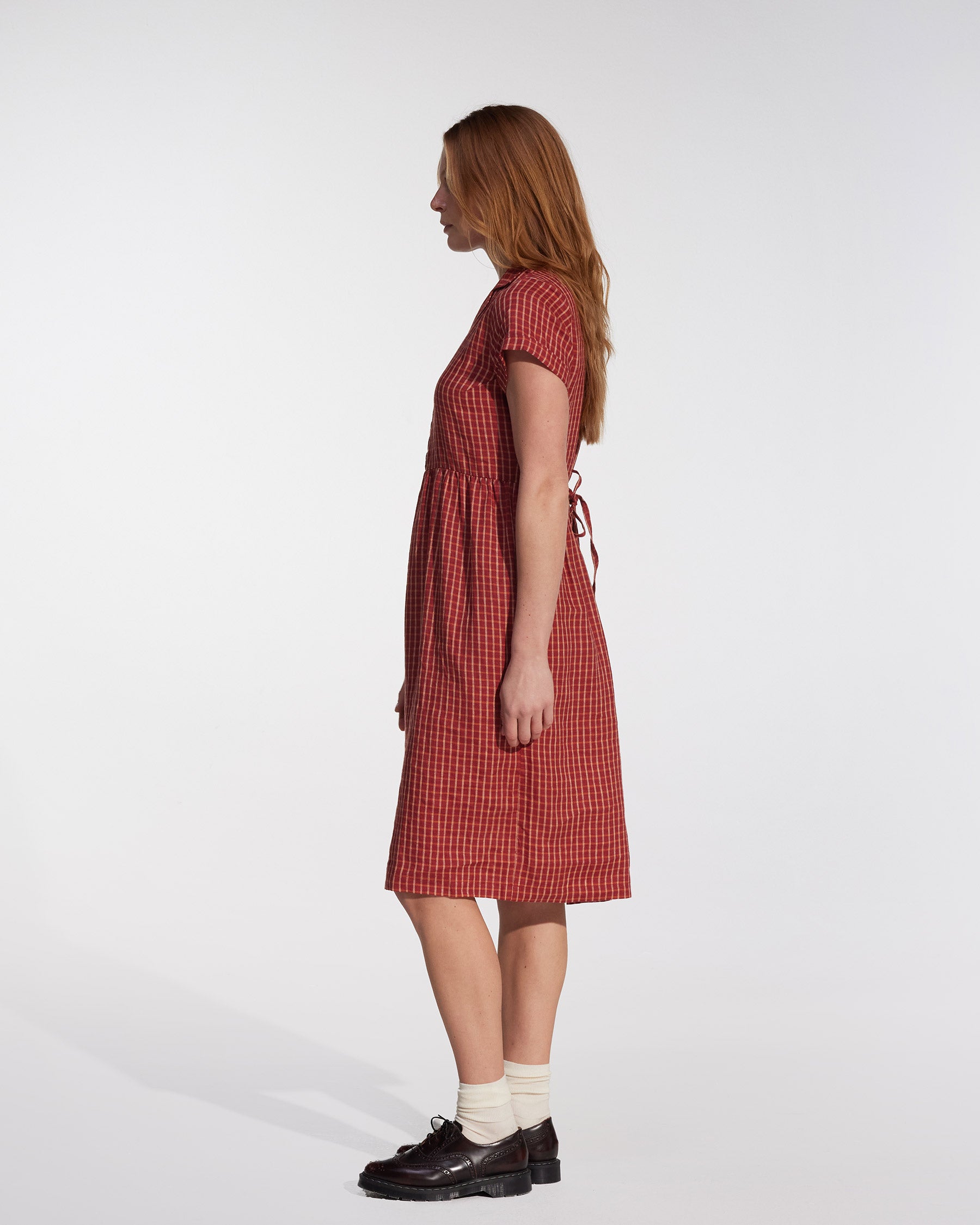 A woman with long light brown hair stands in profile wearing the No.30 Collared Linen Dress in Pomodoro Plaid by Pyne and Smith Clothiers, styled with white socks and dark brown shoes against a plain light background.